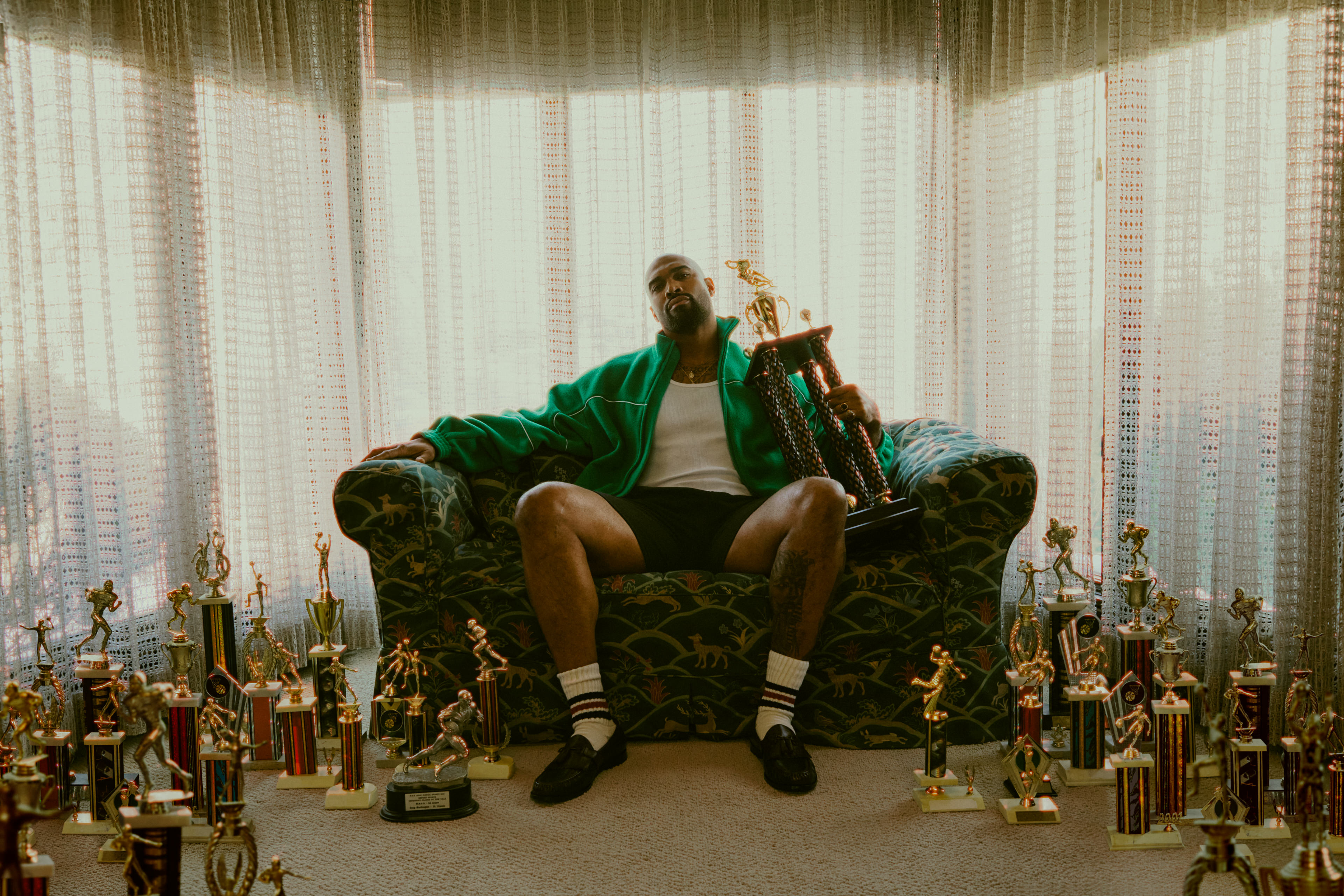 A man in a green jacket sits in an ornate chair surrounded by dozens of football trophies, holding one aloft, backlit by sheer curtains.