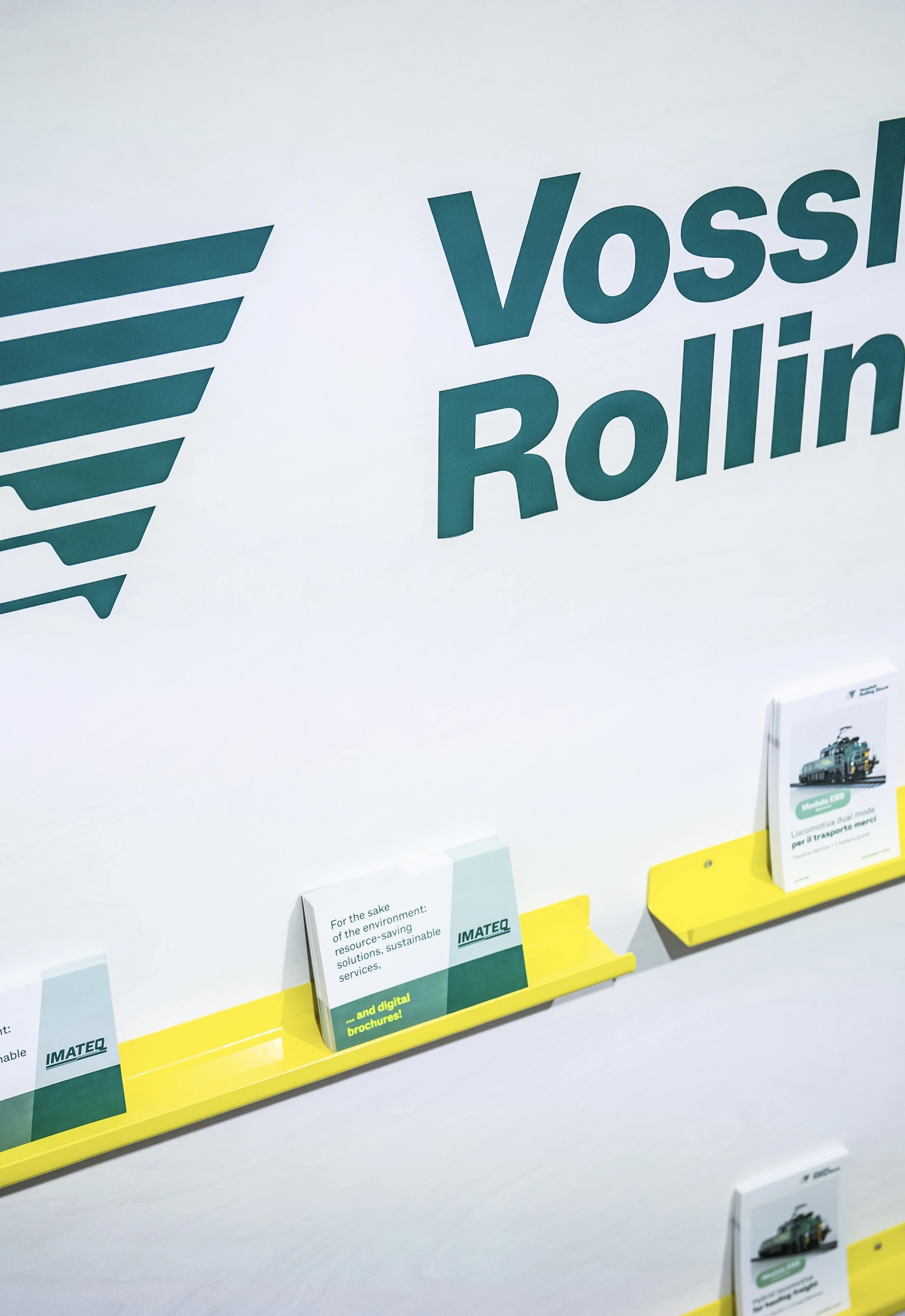 Vossloh Rolling Stock logo wall with yellow shelves holding Imateq brochures at the trade fair booth.