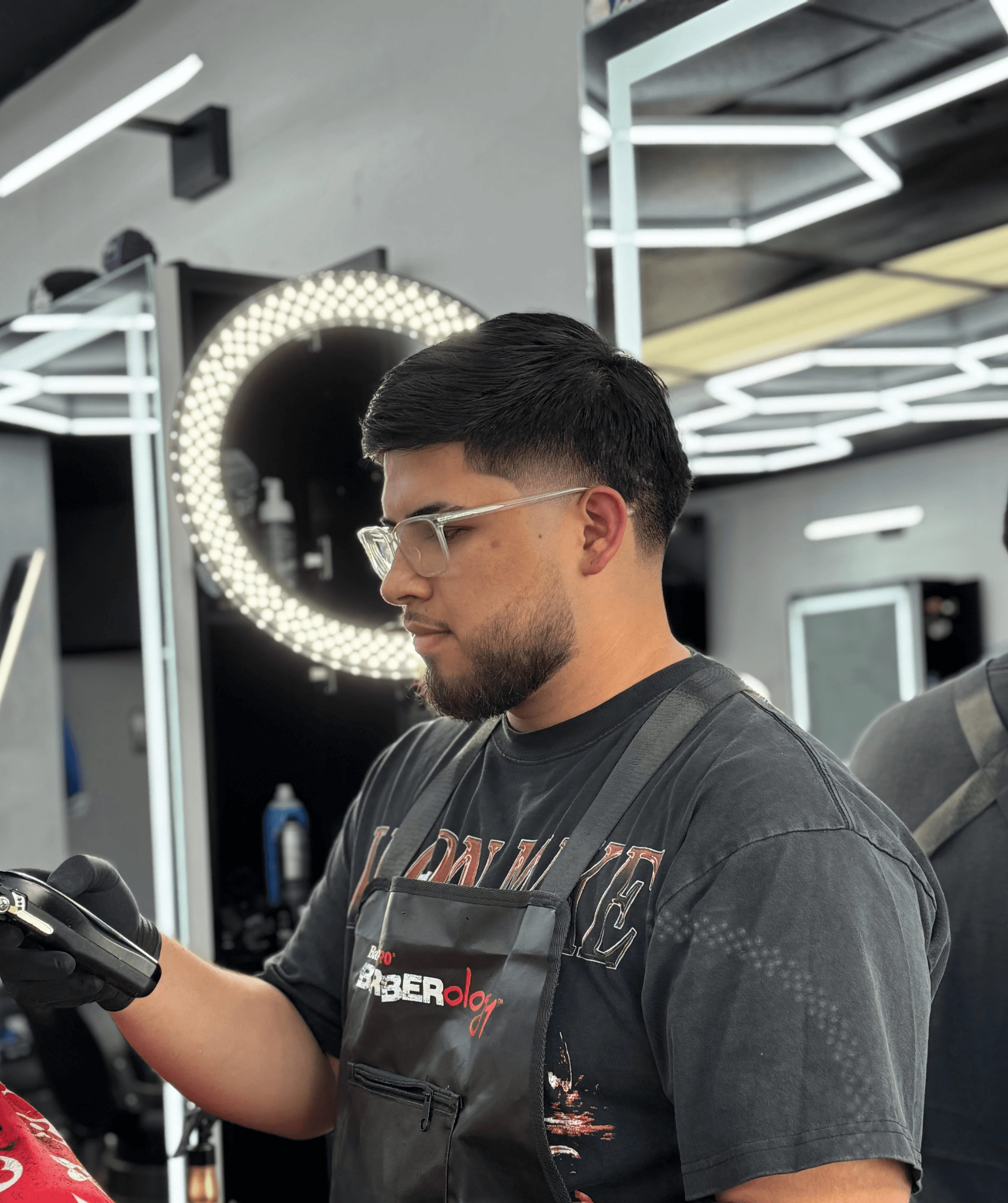 Barber standing with arms crossed in a modern barbershop, wearing a black cap, black T-shirt, and white pants, with hexagon ceiling lights in the background.