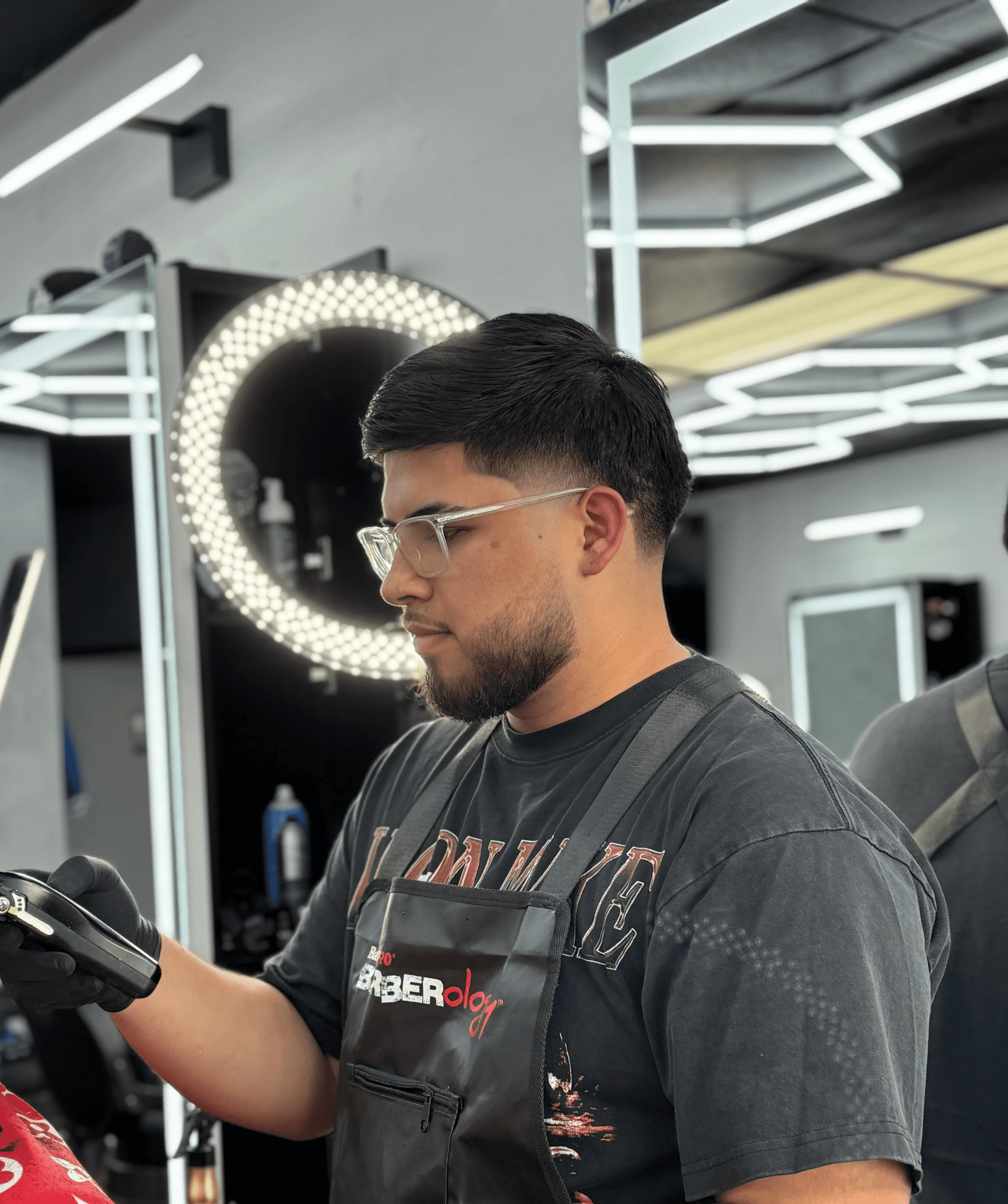 Barber standing with arms crossed in a modern barbershop, wearing a black cap, black T-shirt, and white pants, with hexagon ceiling lights in the background.