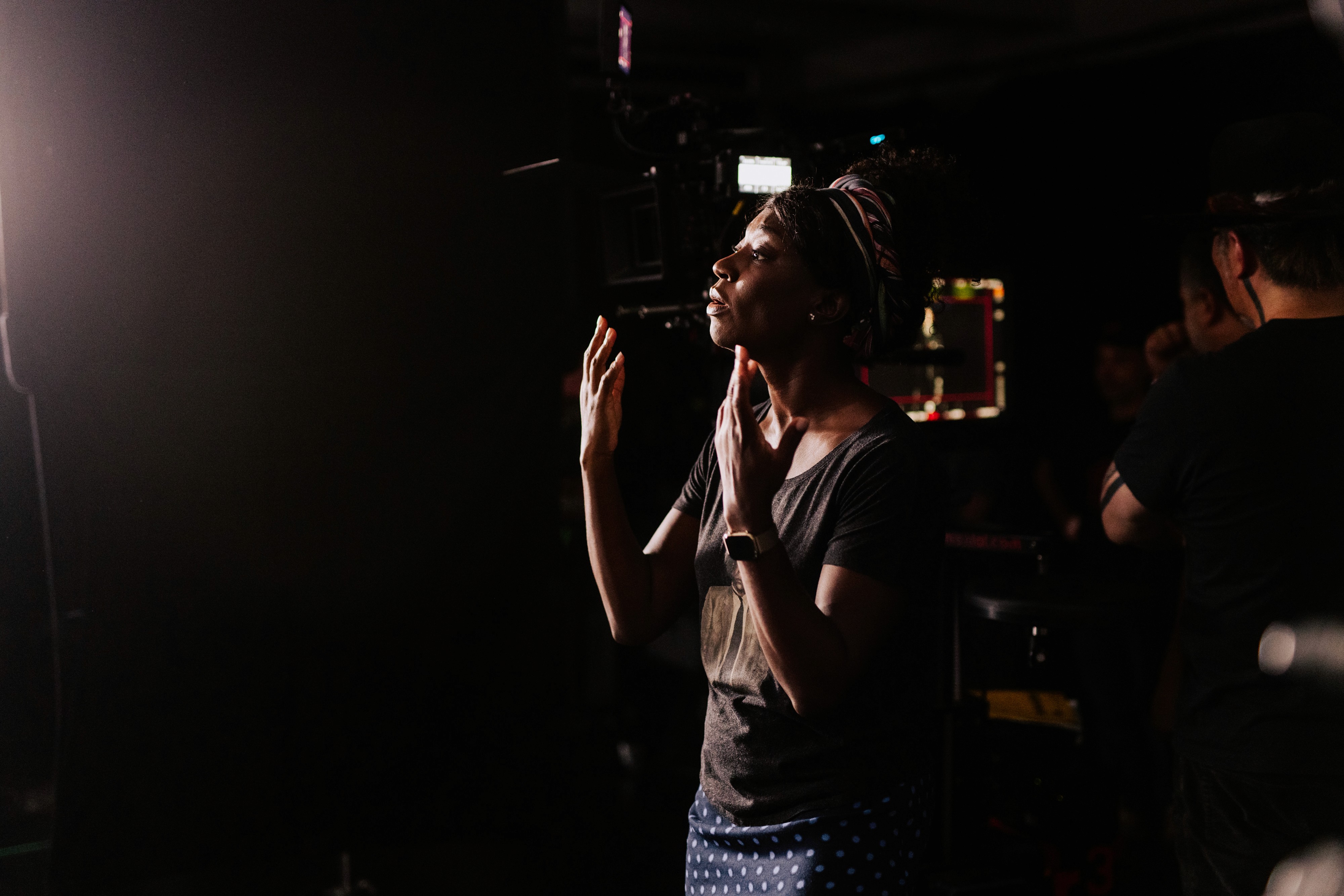 Natasha Ofili (a dark-skinned Black woman) in a dark room, illuminated by soft light, with her hands raised as she teaches Chris Martin the ASL translation.