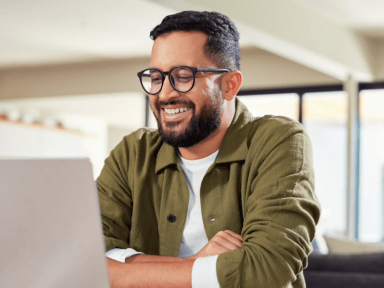 A smiling man with glasses and a beard wearing a green jacket, working at a laptop in an office or café setting