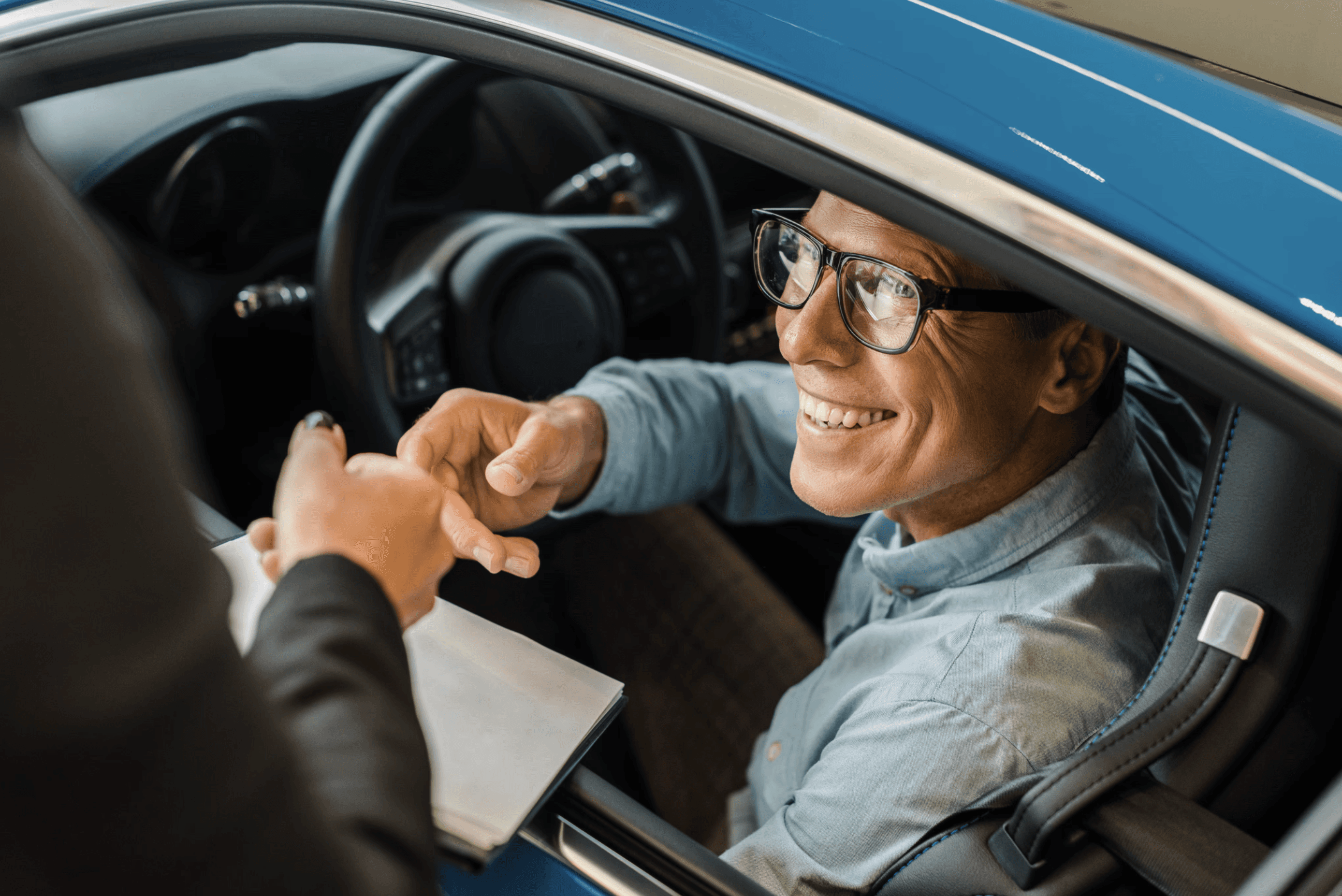 Smiling man sitting in his newly purchased car, receiving keys and paperwork from a dealership representative.