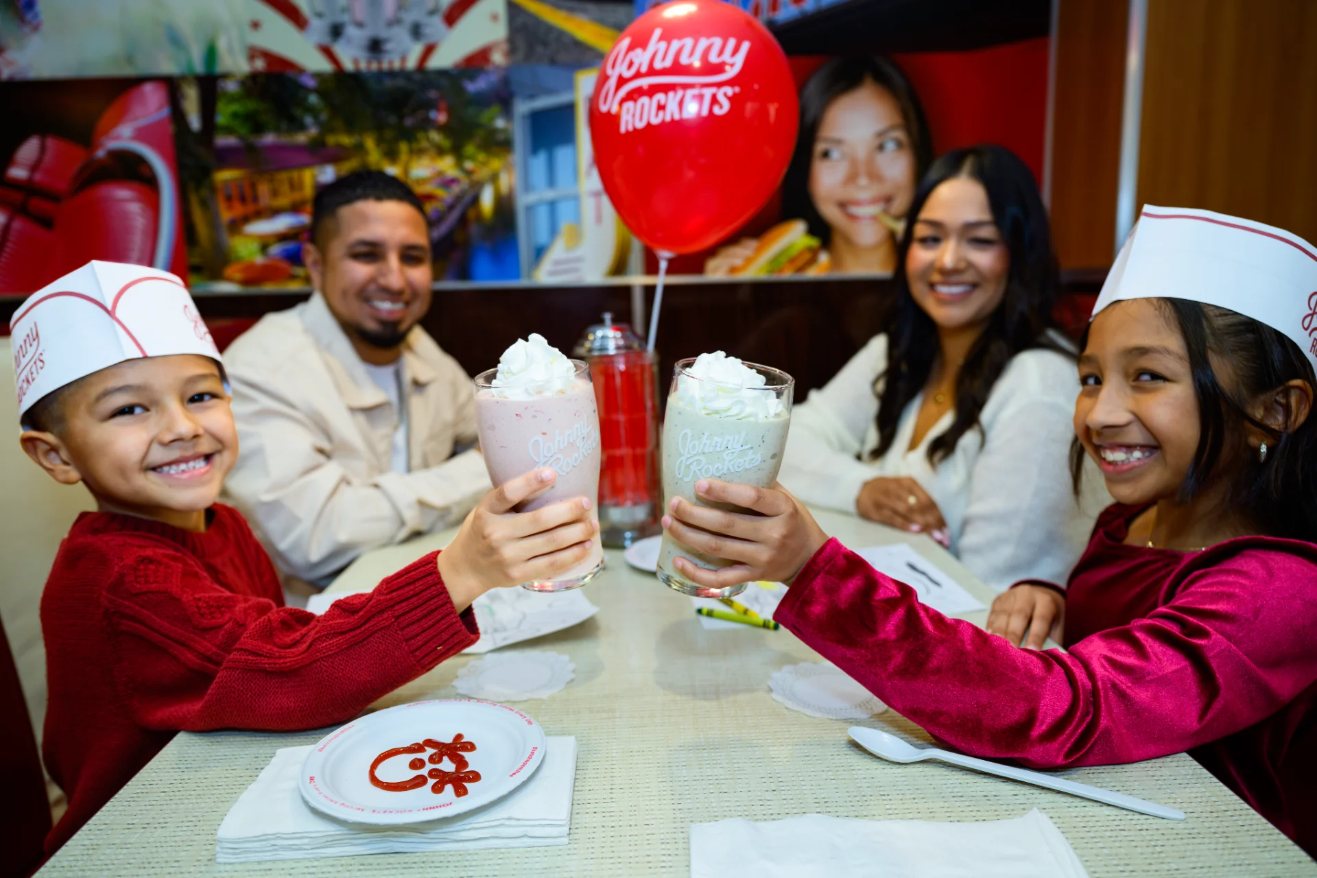 Family in paper hats toasting milkshakes in a Johnny Rockets booth
