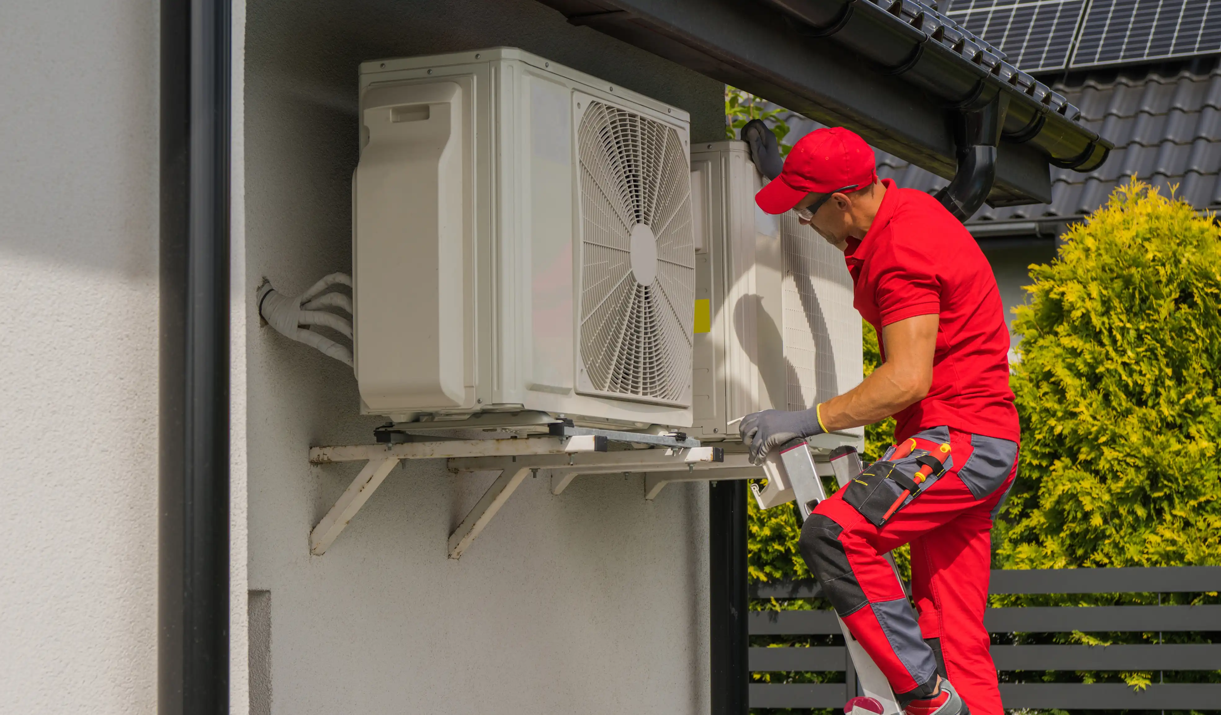 HVAC technician in a red uniform and cap stands on a ladder servicing an outdoor heat pump condenser unit.