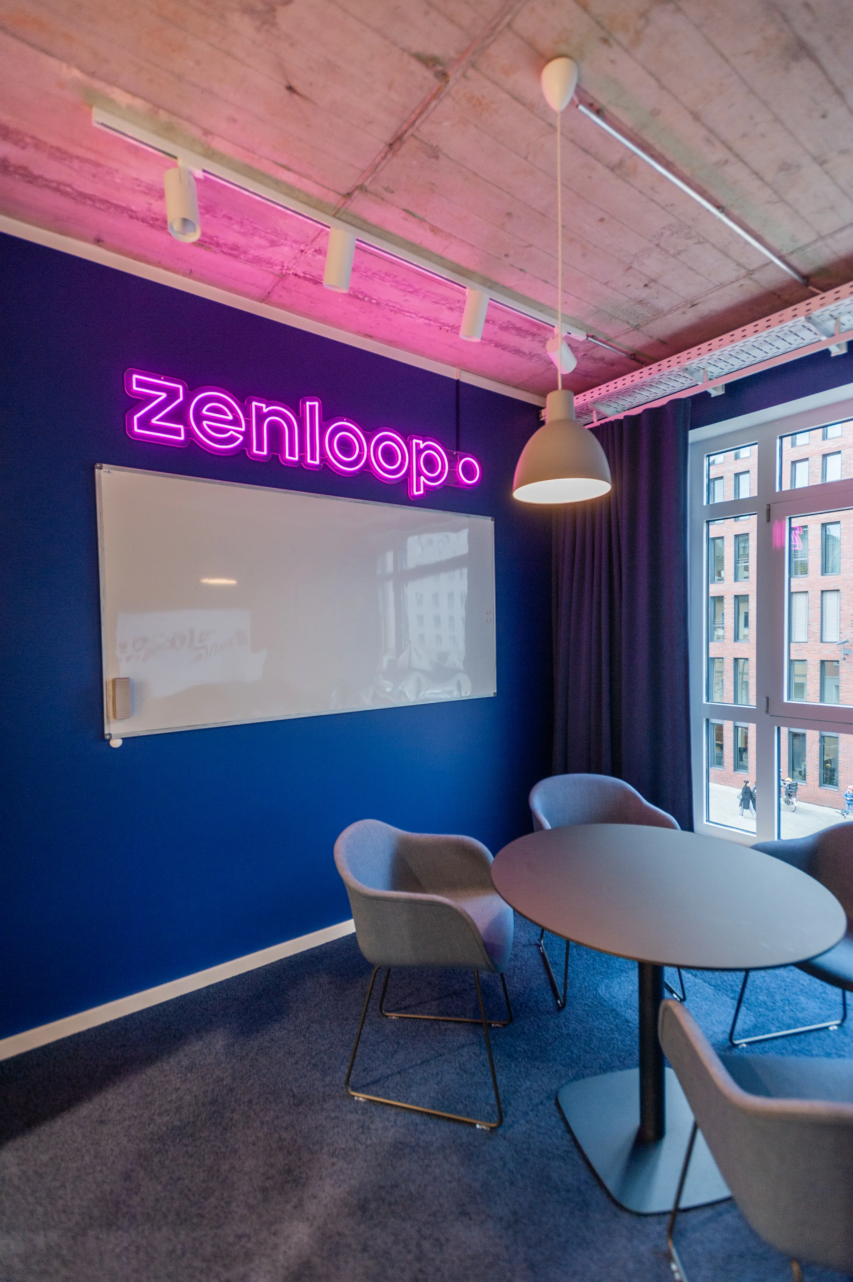 Meeting room at ZENLOOP with a pink zenloop neon sign and whiteboard on a navy wall, a round dark table with grey upholstered chairs, blue carpet and exposed concrete ceiling.