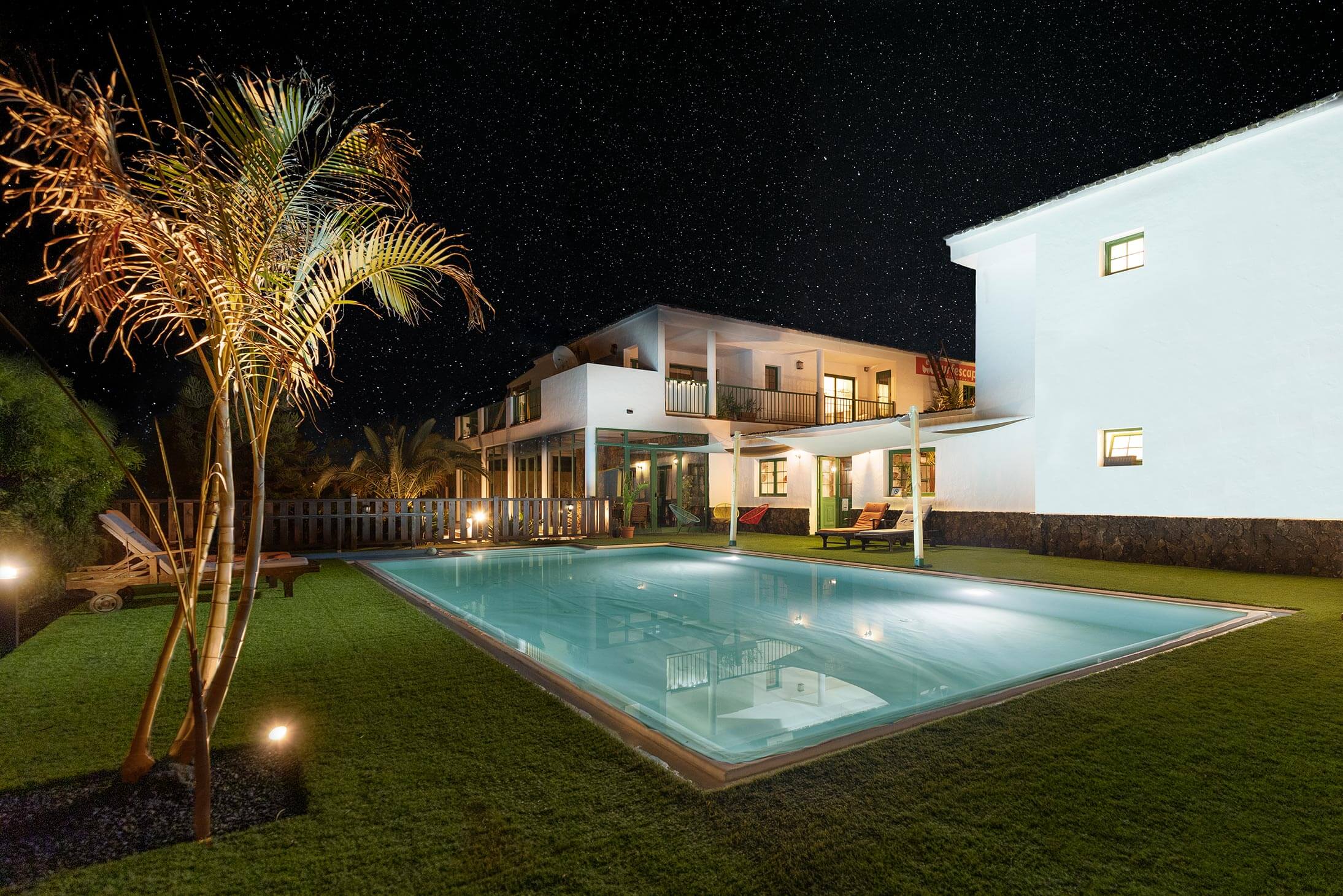 Illuminated outdoor swimming pool at night under a starry sky, surrounded by white Mediterranean villas and palm trees in Fuerteventura.