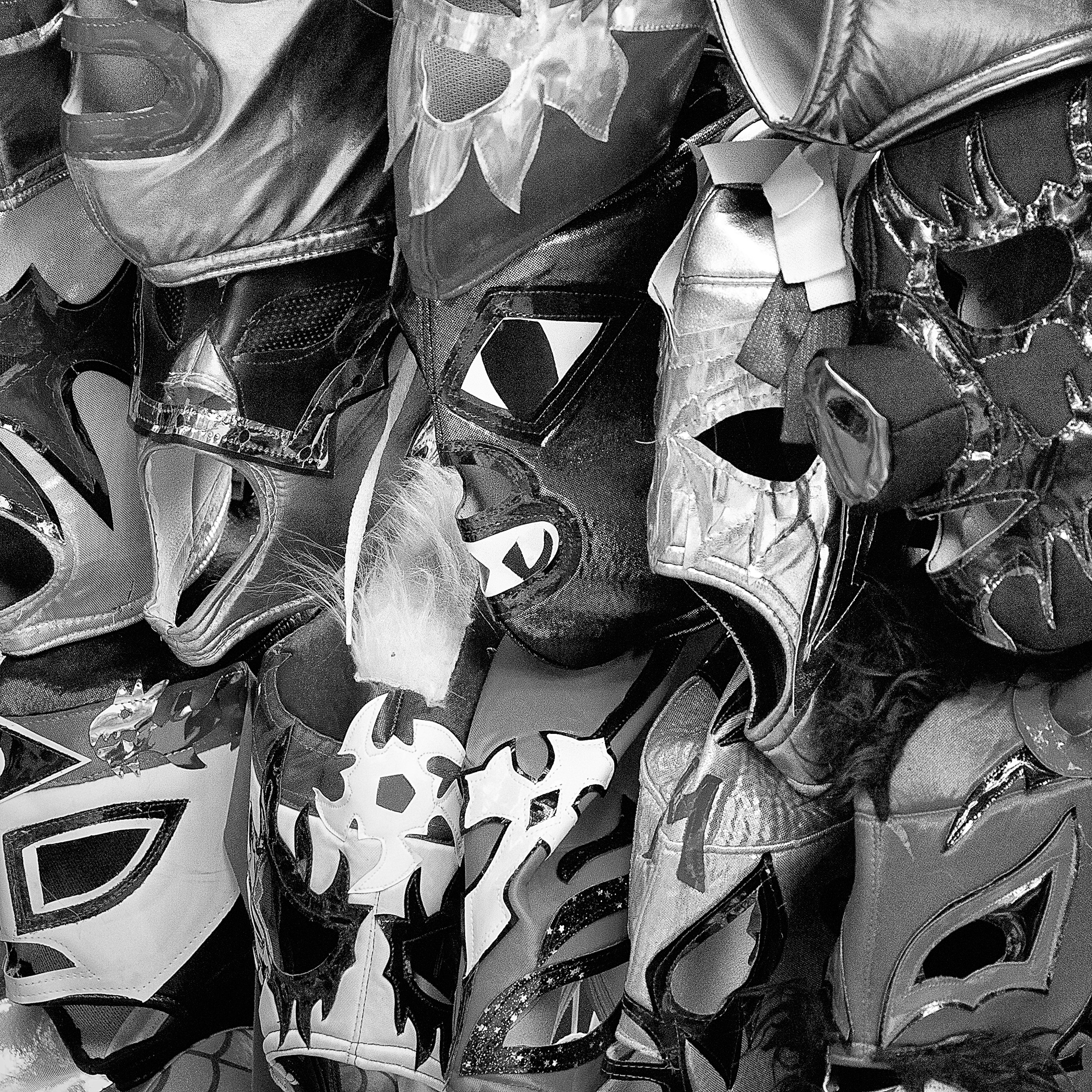 Black and white close-up photo of a dense cluster of lucha libre masks, showcasing a variety of shapes, textures, and dramatic designs.