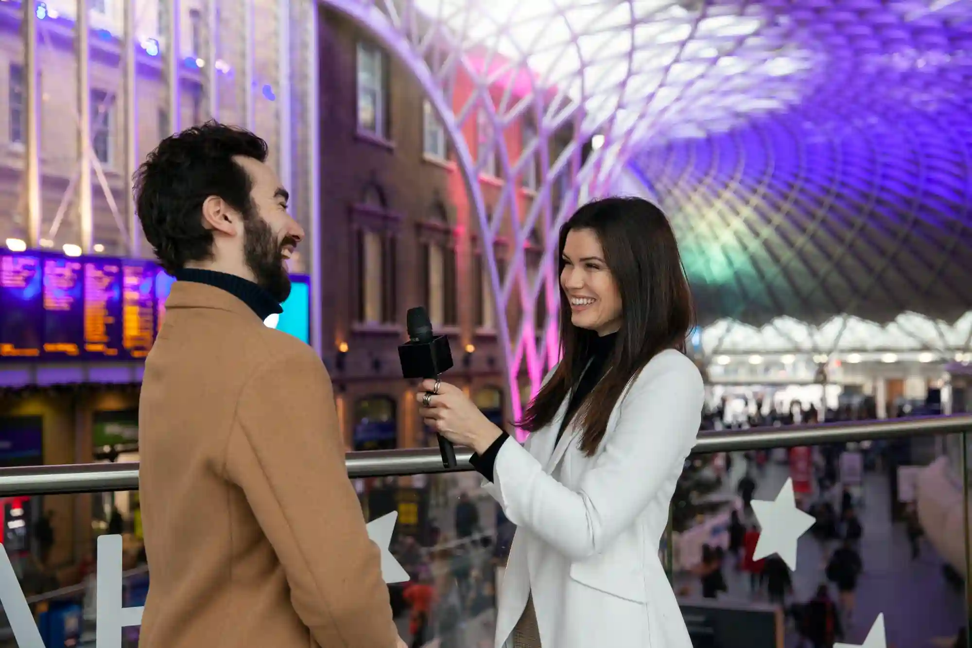 A female journalist interviews a man with a microphone in a busy, modern station for a news segment.