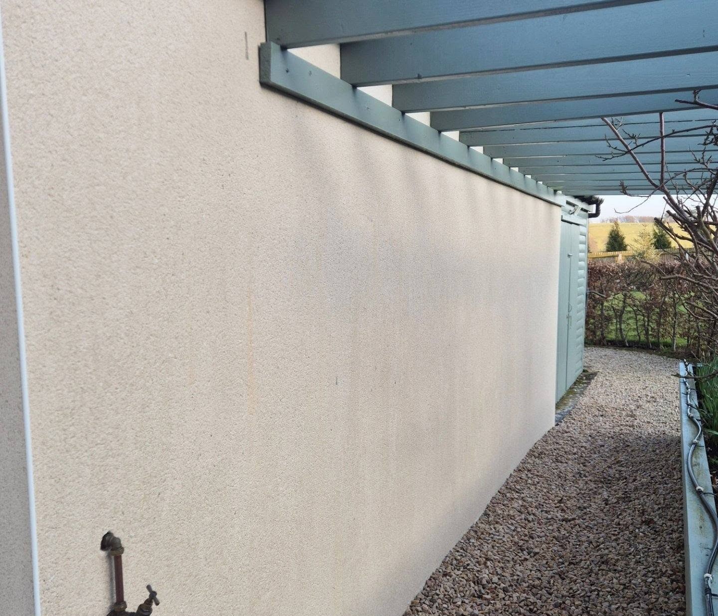 A gravel pathway runs alongside a beige wall under a blue pergola.