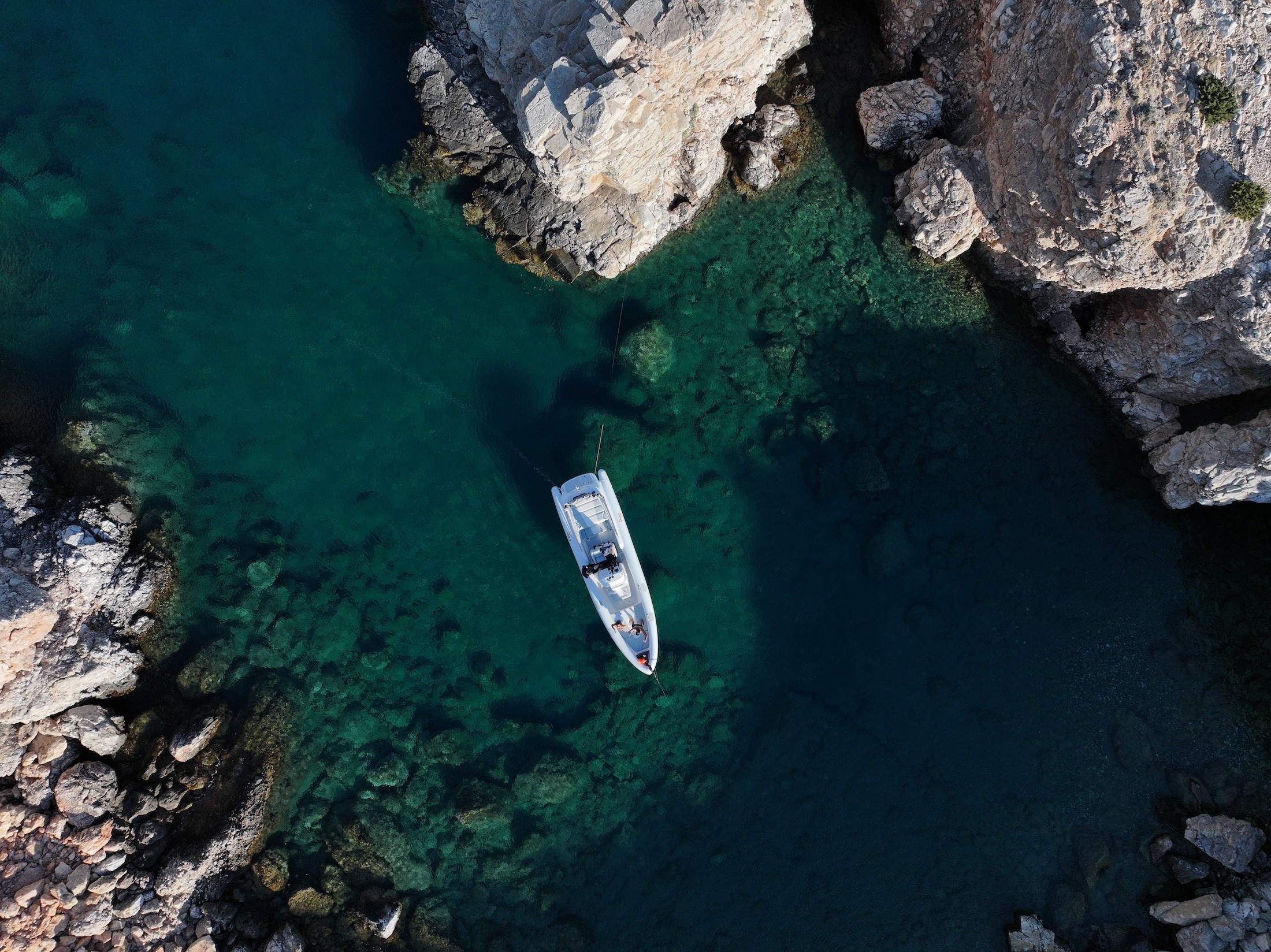 White speedboat with black canopy anchored in crystal-clear turquoise waters, viewed from above showing the pristine Aegean Sea floor beneath.