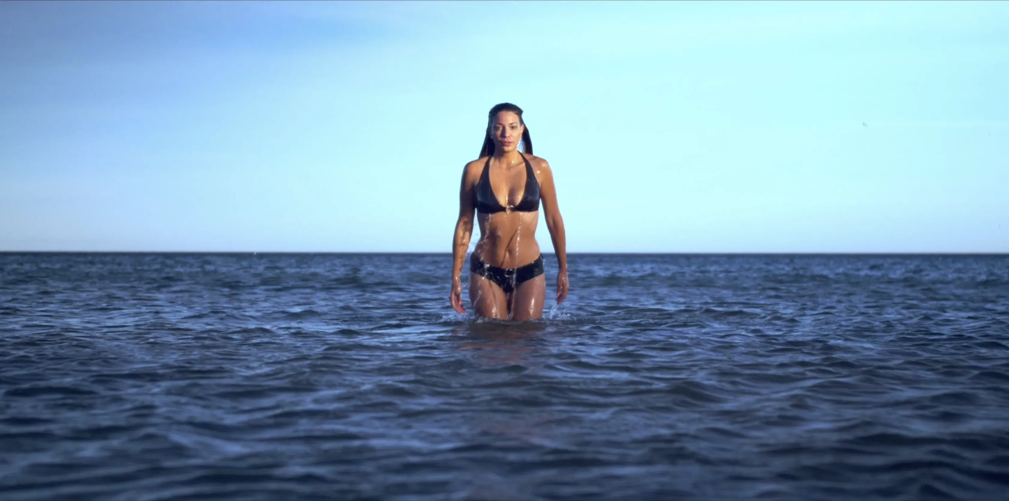 A person in a black bikini emerges from the calm ocean against a clear blue sky, showcasing a serene beach setting.