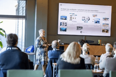 A presenter speaking to an audience in a conference room, with a large screen displaying images and information.