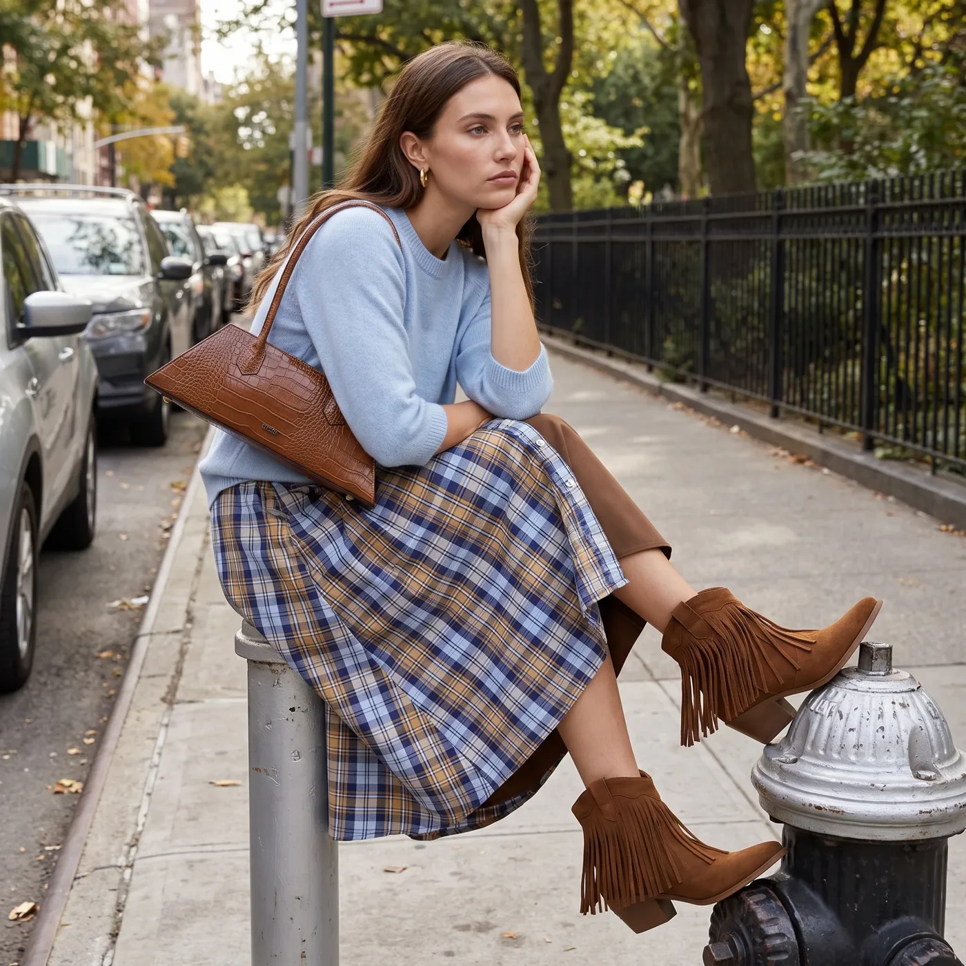 Ragazza seduta in strada con maglione azzurro, gonna a quadri, borsa effetto cocco e stivaletti con frange