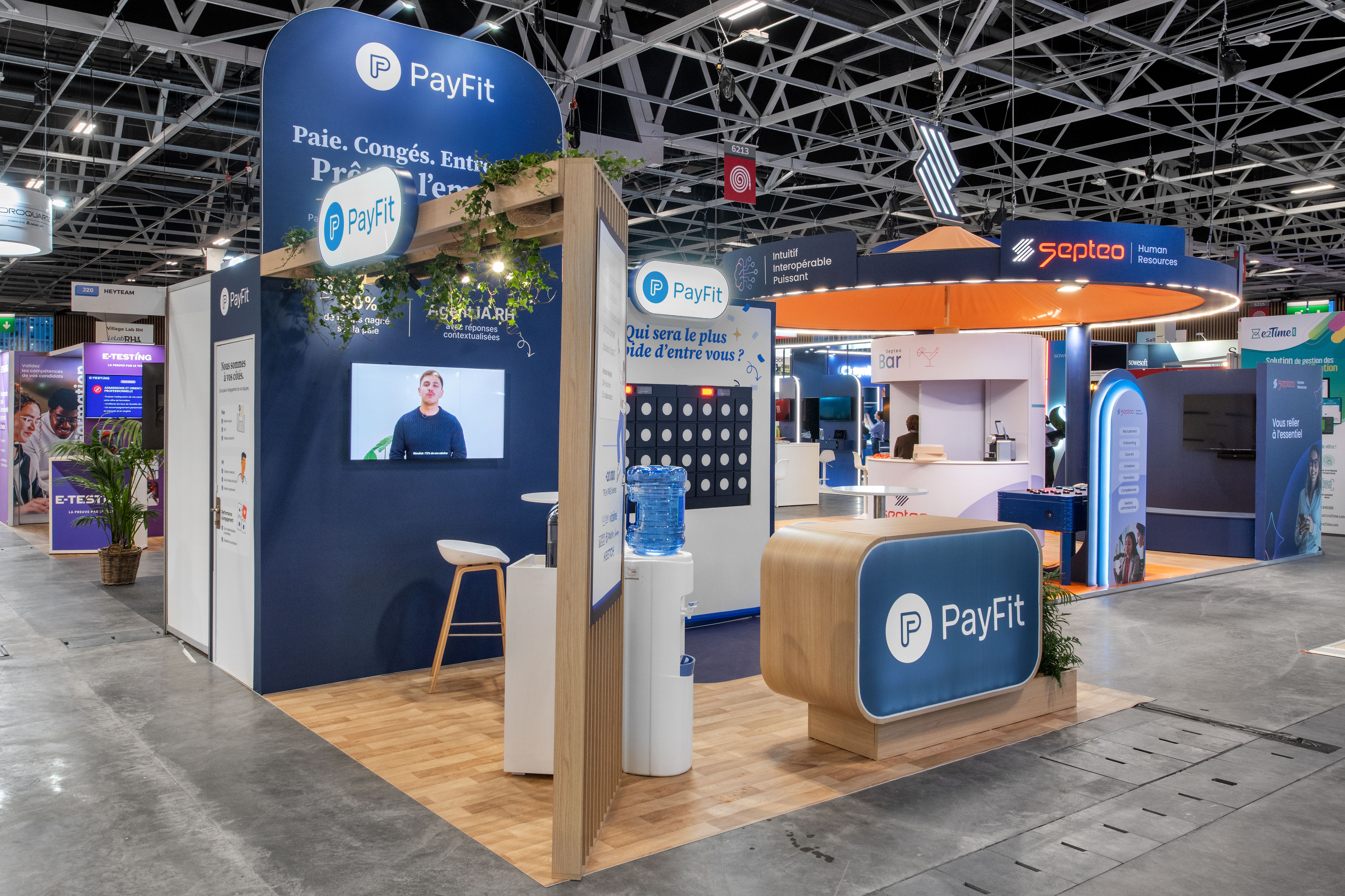 Stand PayFit salon HR Technologies 2026 réalisé par Expace – vue frontale avec arche bois végétalisée, fond bleu marine Agent IA RH, jeu interactif Puissance 4 et comptoir d'accueil arrondi bois et bleu
