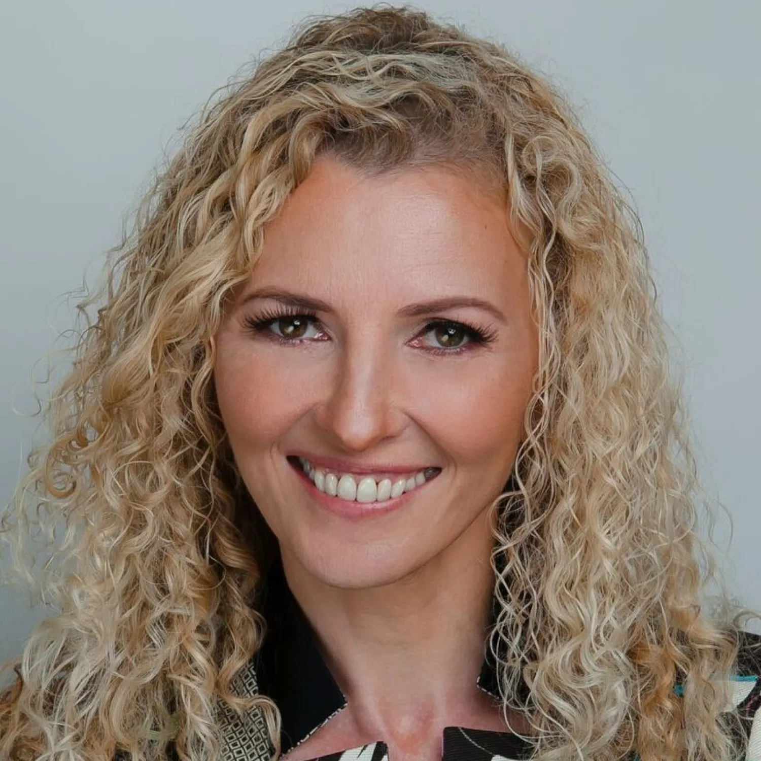 A young person with curly hair smiles at the camera while sitting in front of a laptop against a dark background.