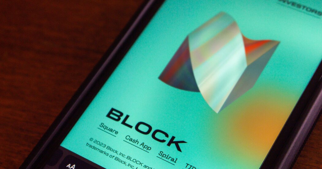 Nahaufnahme eines Smartphone-Bildschirms mit türkisfarbener 3D-Form und dem Schriftzug „BLOCK“ samt zugehöriger Unternehmen, auf einer Holzoberfläche.