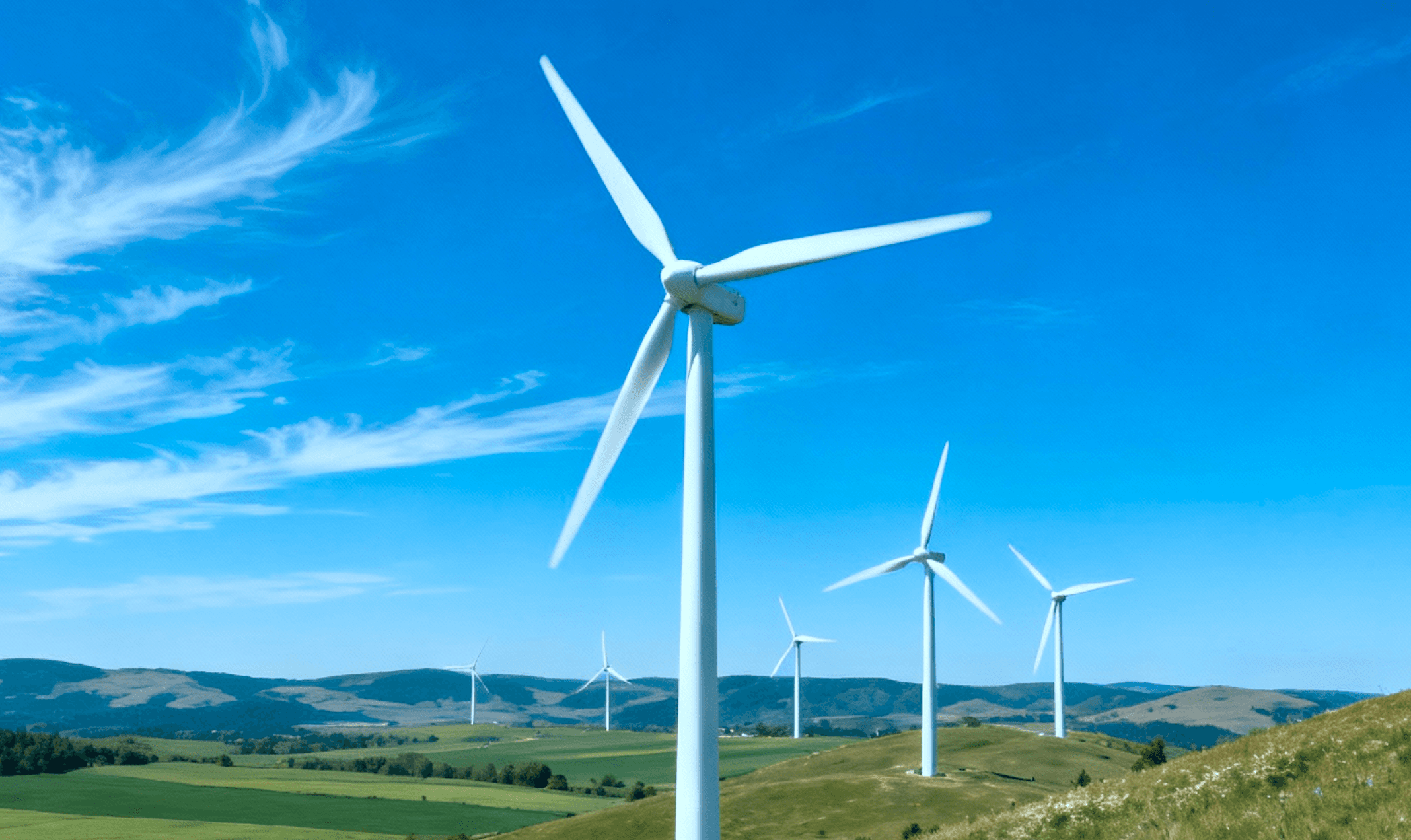 Wind turbines are positioned on a grassy hill under a blue sky with scattered clouds, generating renewable energy.