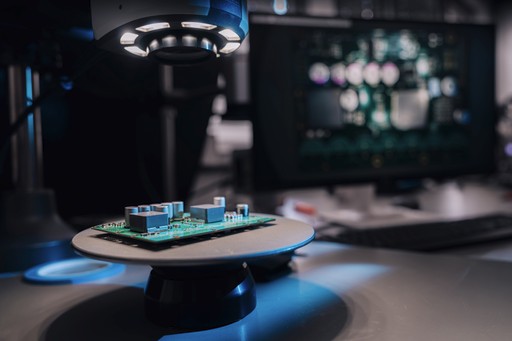 A circuit board on a raised platform, illuminated by a top light on a desk, with blurry screens in the dark background.
