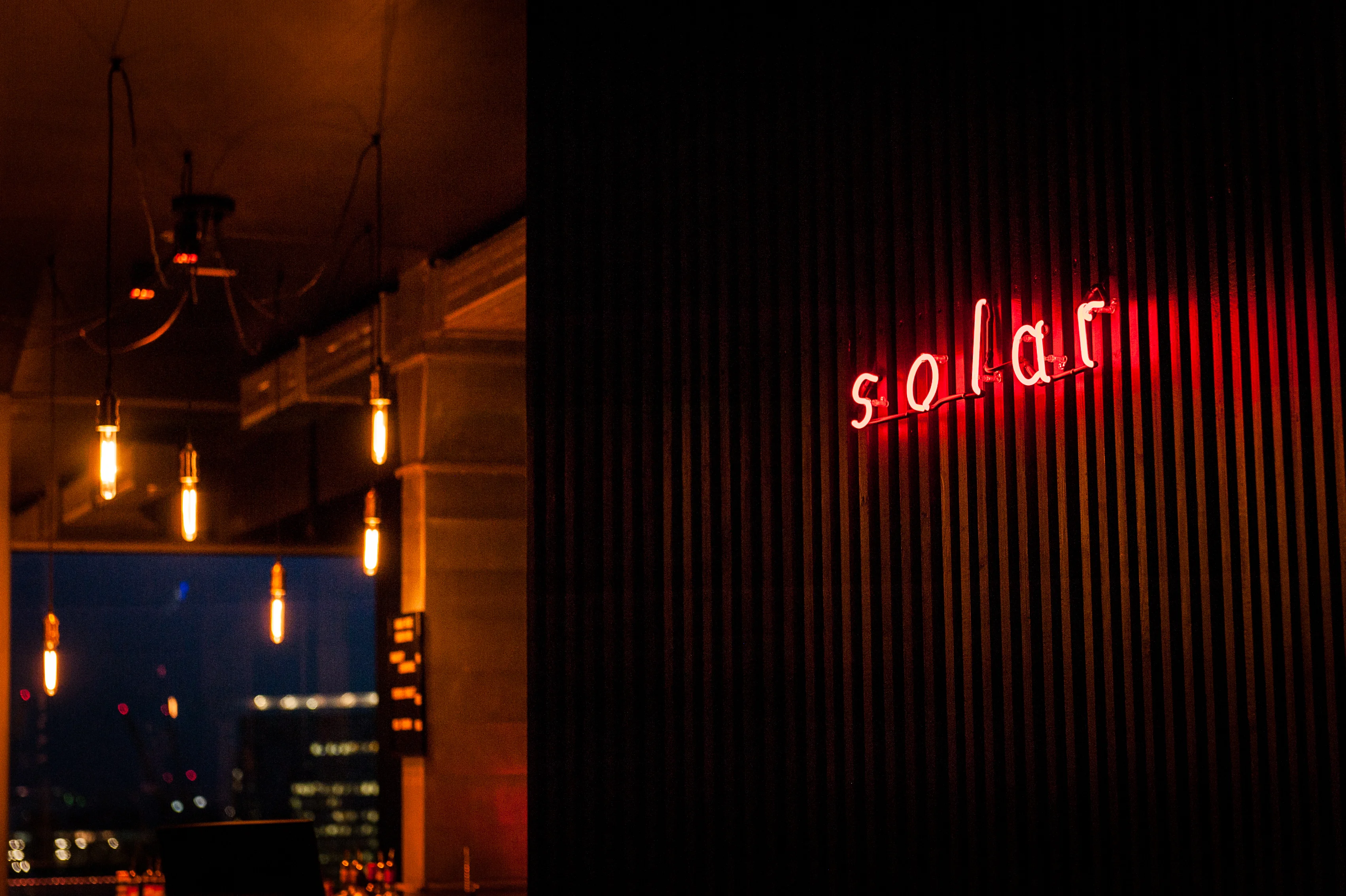Red neon sign reading solar on a dark wooden slat wall at the entrance of SOLAR SKY LOUNGE, with warm filament bulbs and a night city view to the left.