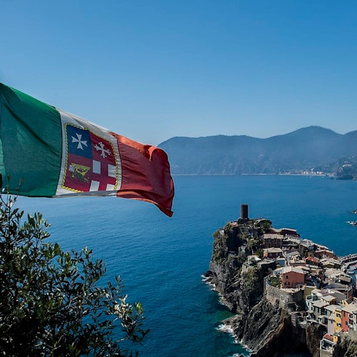 一座海边村庄坐落在悬崖边，房屋色彩缤纷，前景有一面旗帜，背景是连绵群山。