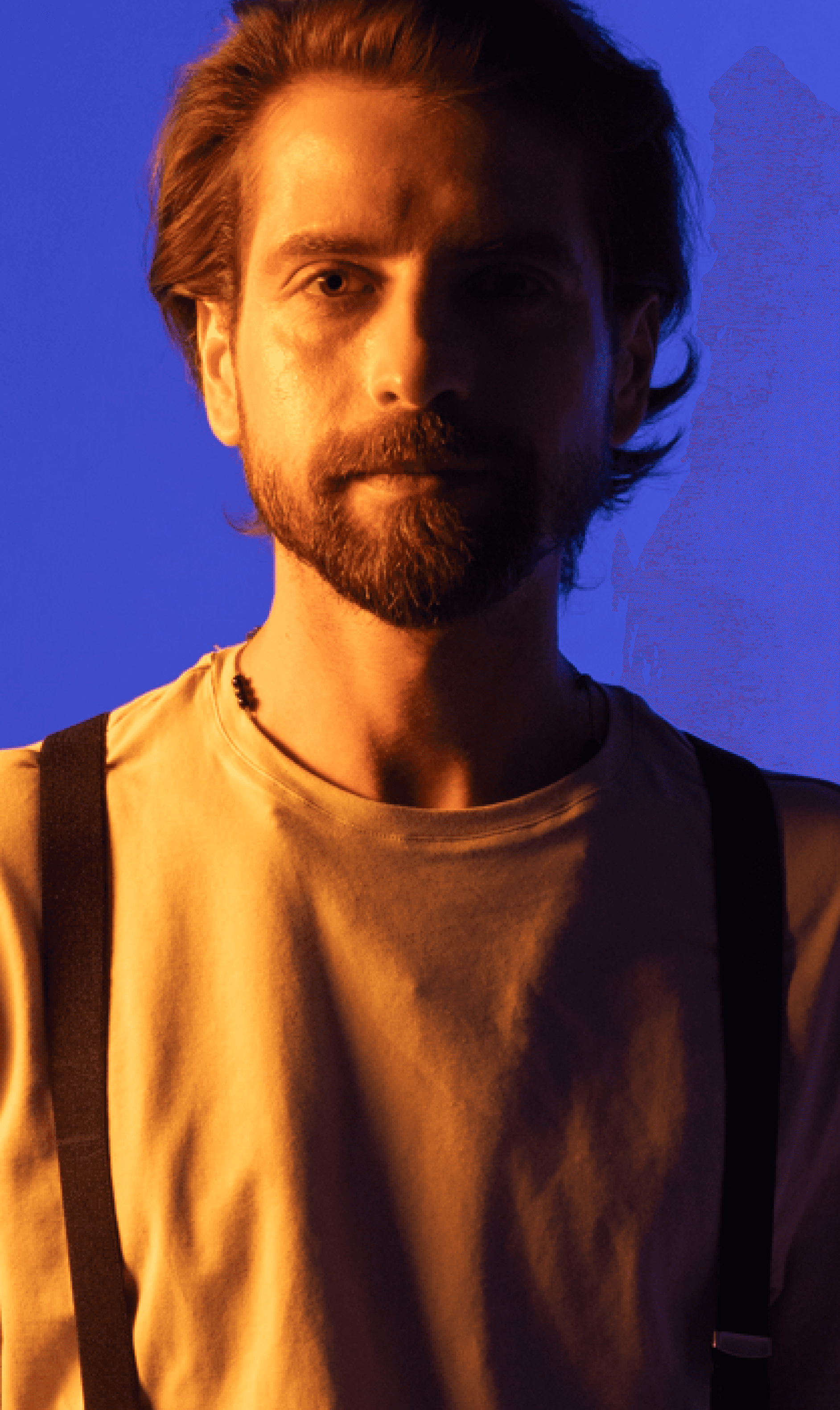 Pełen wyrazu portret brodatego muzyka zespołu Electric Shapes patrzącego przed siebie, ubranego w jasną koszulę i szelki, z wyraźnym kontrastem między ciepłym oświetleniem twarzy a chłodnym niebieskim tłem.