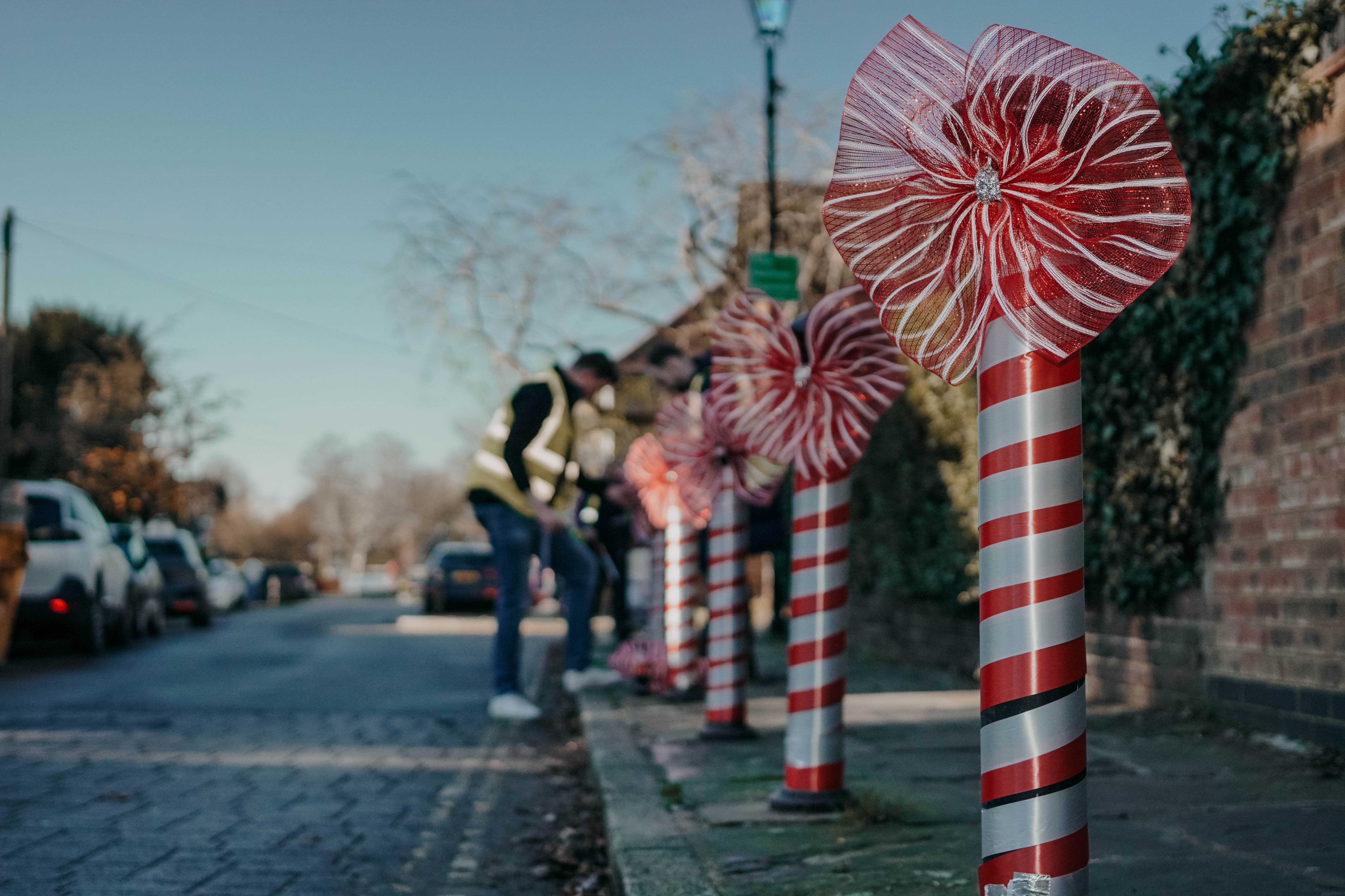 Large, festive candy cane decorations with red and silver stripes line a street curb on a sunny day. In the foreground, one decoration is topped with a large red bow, while in the background, a person sets up more decorations.