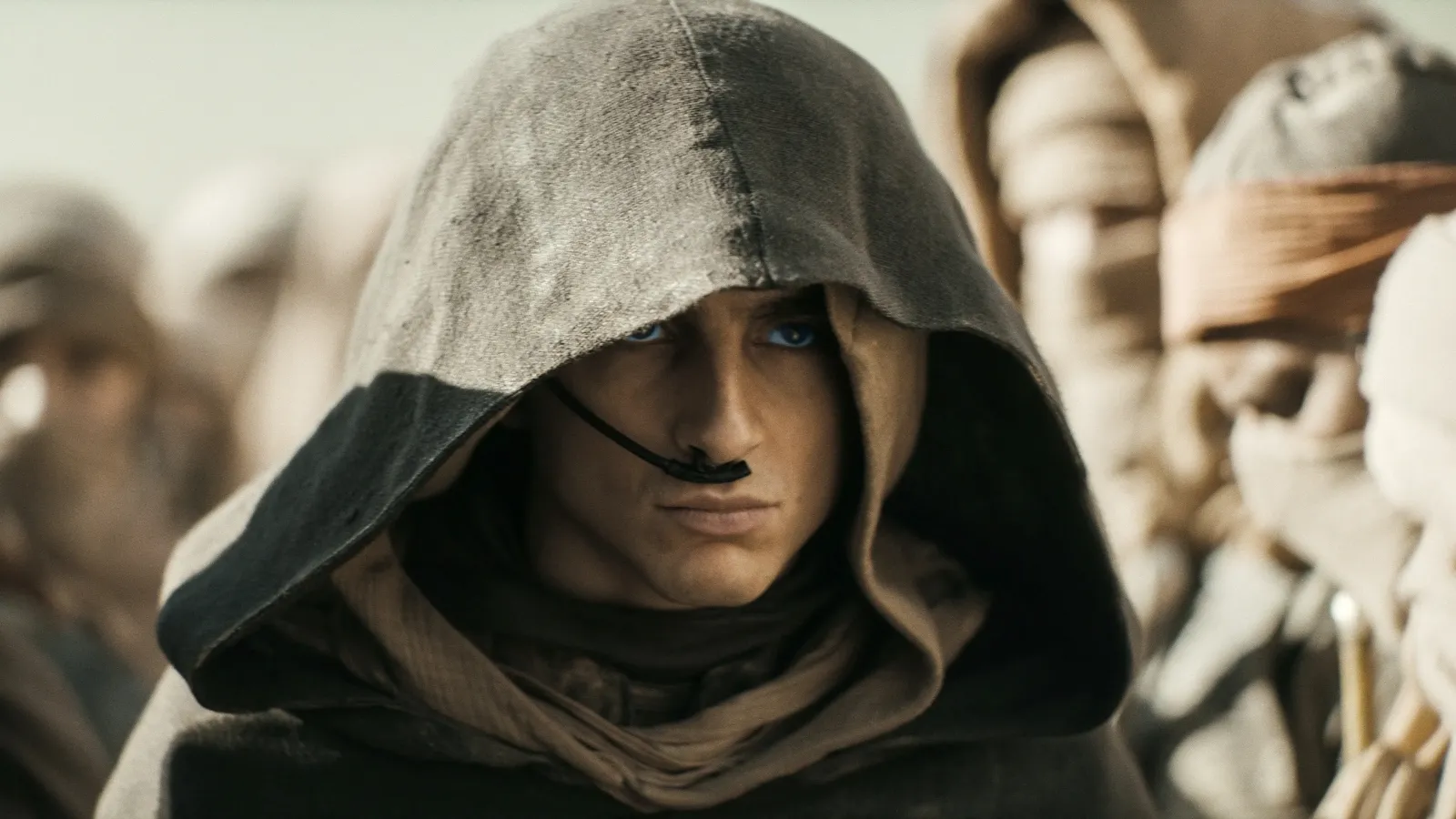 A young man wearing a hooded cloak and scarf with vibrant blue eyes stands among a group of similarly dressed individuals in a desert setting.