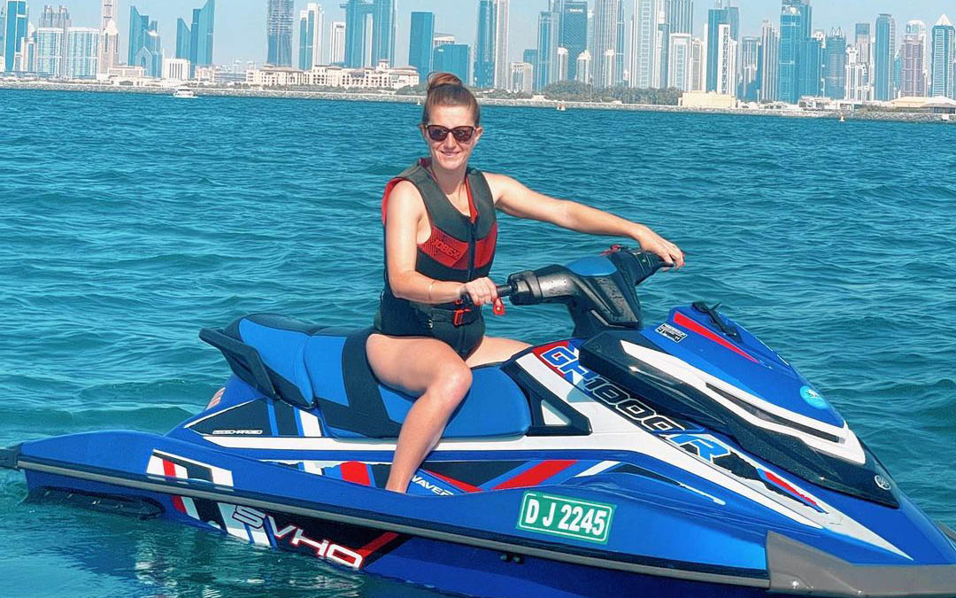 A happy woman on a blue jet ski in Dubai, wearing a life jacket and sunglasses, showcasing a safe and practical outfit for jet ski adventures, highlighting what to wear for jet skiing, with the city skyline in the background on a sunny day on the water