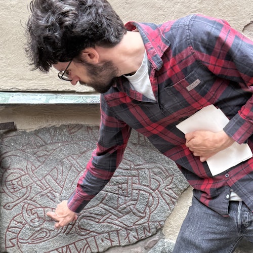 A man in a red plaid shirt points at a stone carving with intricate patterns, holding a white sheet of paper in his other hand.