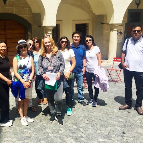 Skupina devíti lidí stojí na dlážděné ulici před obloukovou budovou za slunečného dne.