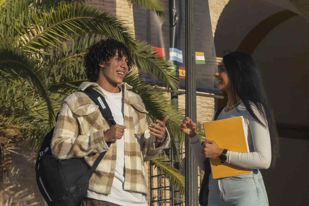 Two students, one wearing a plaid jacket and backpack and the other holding a yellow notebook, engage in conversation outside UCAM University, surrounded by palm trees and colorful flags.