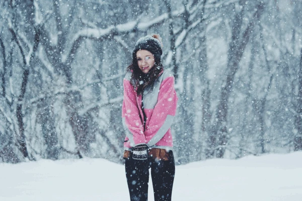 Woman in a pink and gray winter jacket standing in a snowy forest during active snowfall.