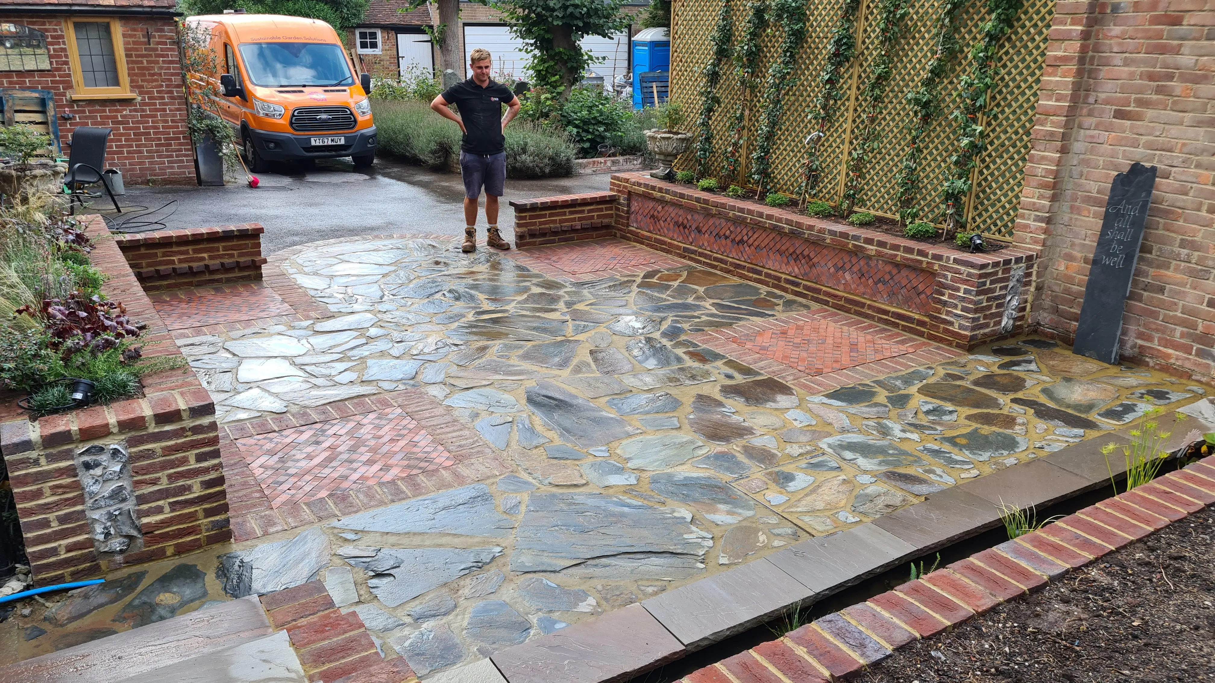 A stone pathway and seating area in a garden with wet stones, surrounded by greenery and a subtle brick wall.