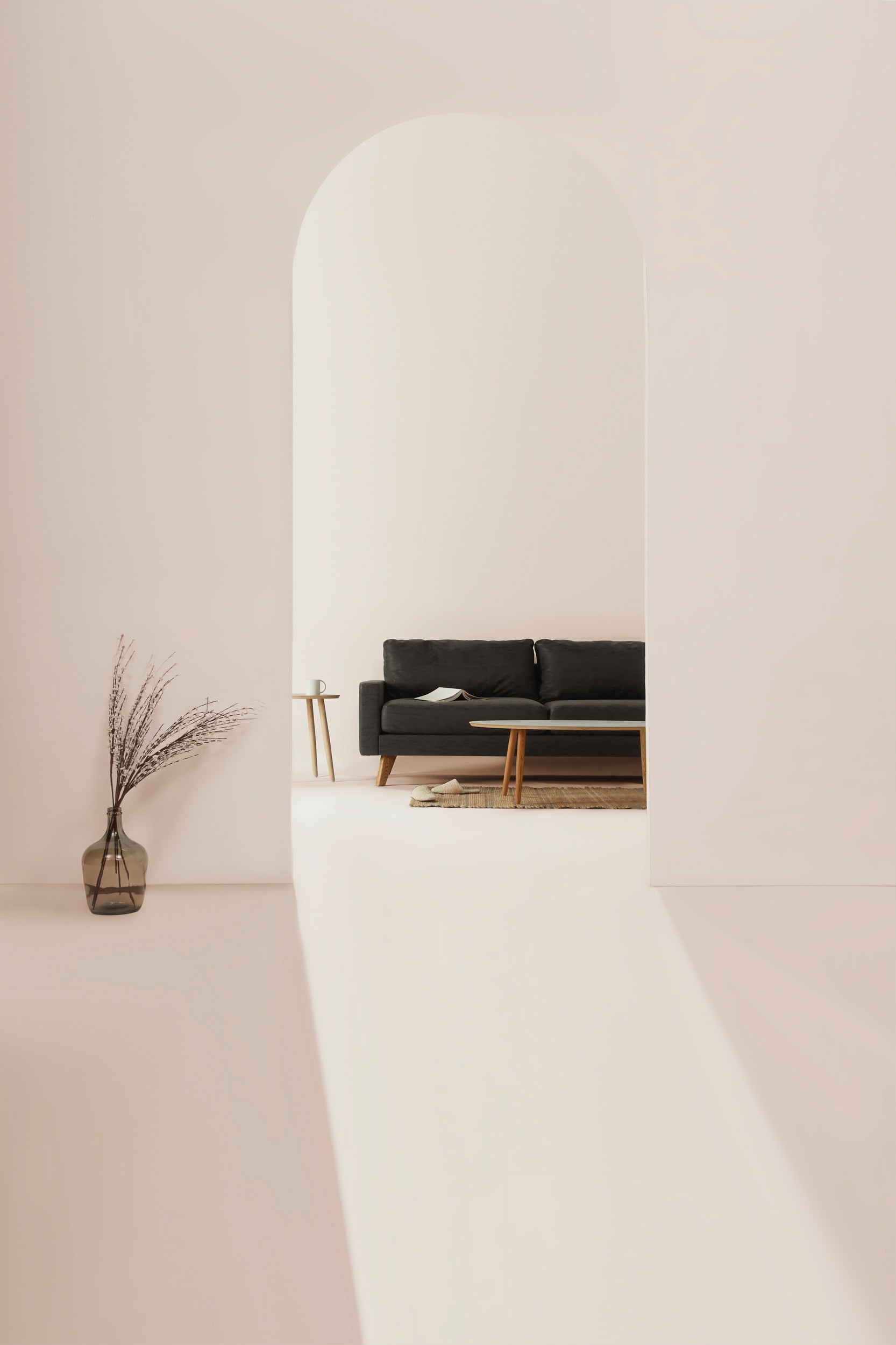Minimalistisches Interieur mit einem sanft beleuchteten, cremefarbenen Bogendurchgang, der den Blick auf eine moderne Wohnzimmerlandschaft mit dunklem Sofa, Holztisch und natürlicher Dekoration lenkt. Ein transparenter Glasvase mit getrockneten Zweigen steht im Vordergrund.