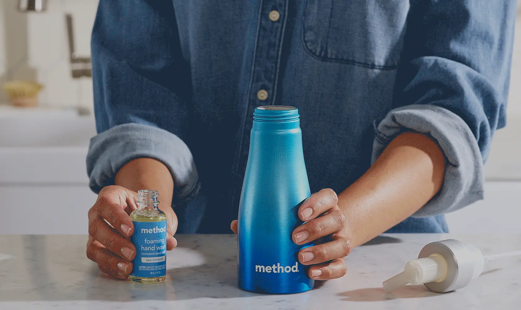 Person pouring a small concentrate vial into a blue aluminum bottle on a kitchen counter, pump cap set beside it