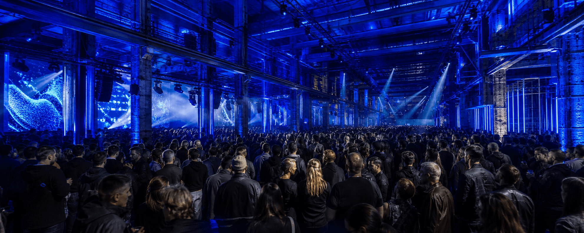 Dense audience inside an industrial creative technology conference hall under cobalt directional lighting