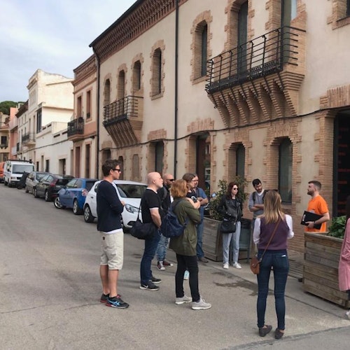 Группа людей стоит на улице перед зданием с арочными окнами и балконами, общаясь друг с другом.