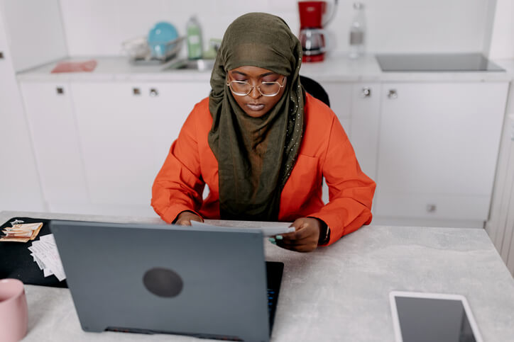 A woman in a hijab sits at a desk, focused on her laptop, with a thoughtful expression on her face. Form G-28