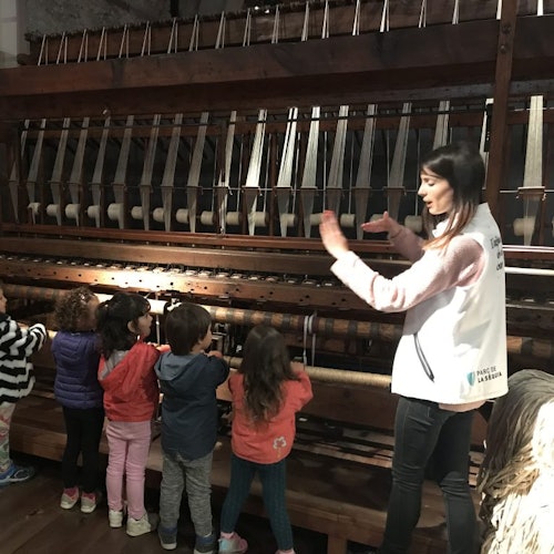 一位女士向五个站在机械织机前的孩子解释如何操作织机，他们正在仔细观察这台机器。