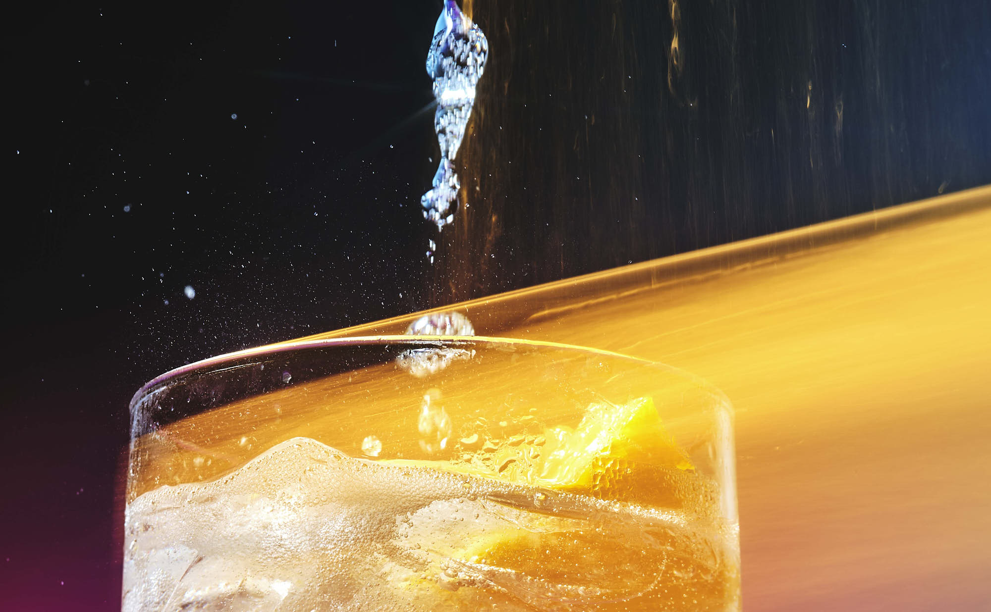 A droplet of liquid hangs above a vibrant orange surface with a dark background, creating a striking visual contrast.