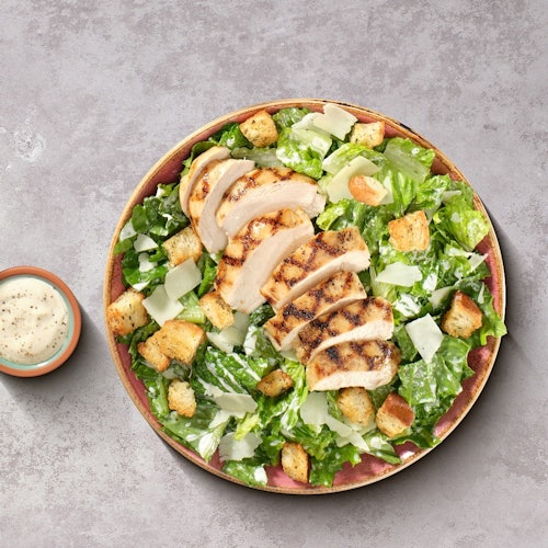 A Caesar salad with grilled chicken slices, croutons, and shaved Parmesan. A small dish of dressing is beside the plate.