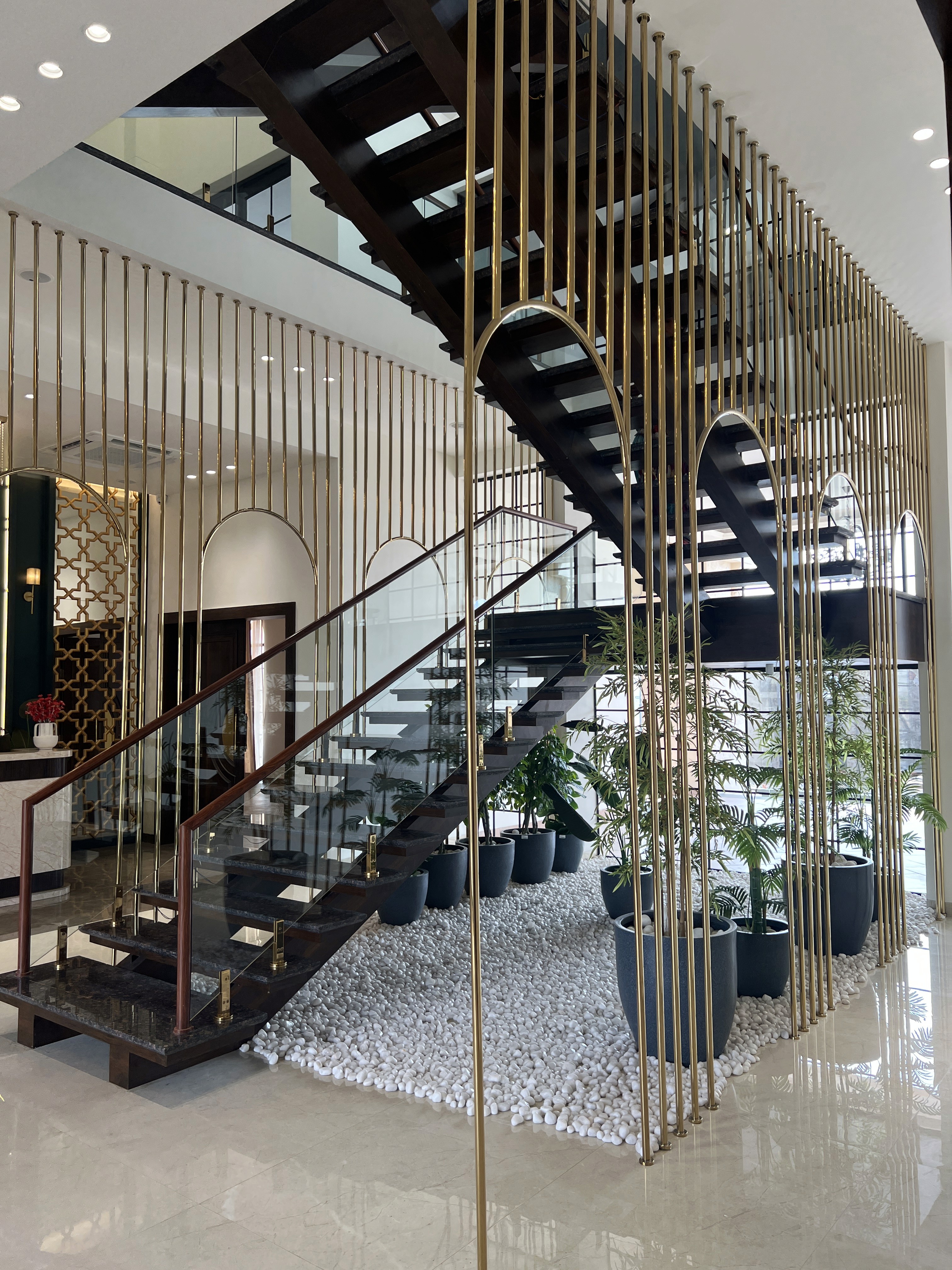 Modern grand staircase design in a commercial lobby featuring dark wooden treads, decorative vertical brass poles, and indoor landscaping with white river rock.