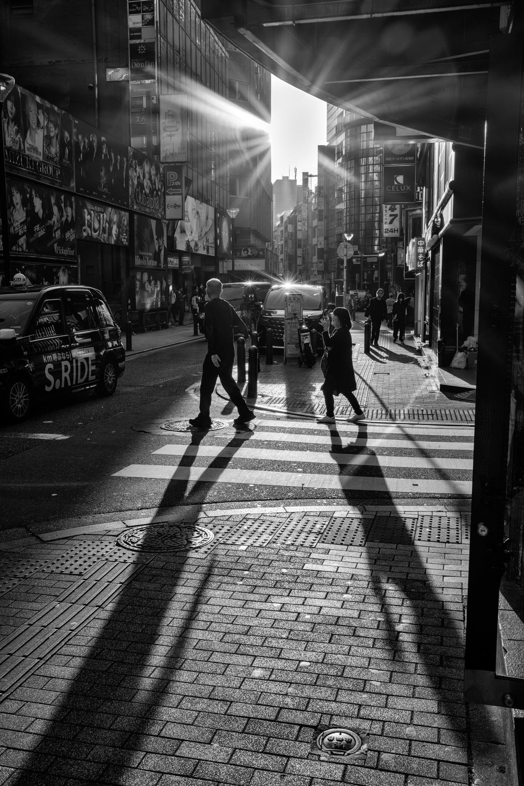 Black and white photograph of pedestrians crossing a street with hard shadows stretching across the pavement.