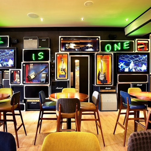 En stilfuld bar med borde, stole og en vægudsmykning med guitarer, forstærkere, neonskilte og tv-skærme.