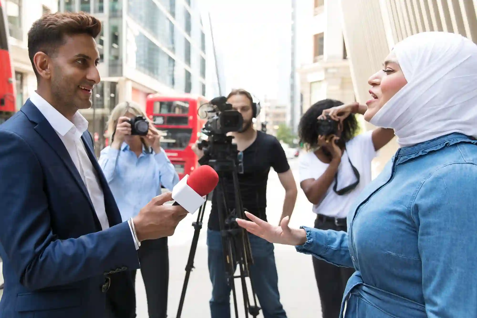 A male reporter interviewing a woman in a hijab on a busy London street with a film crew behind.