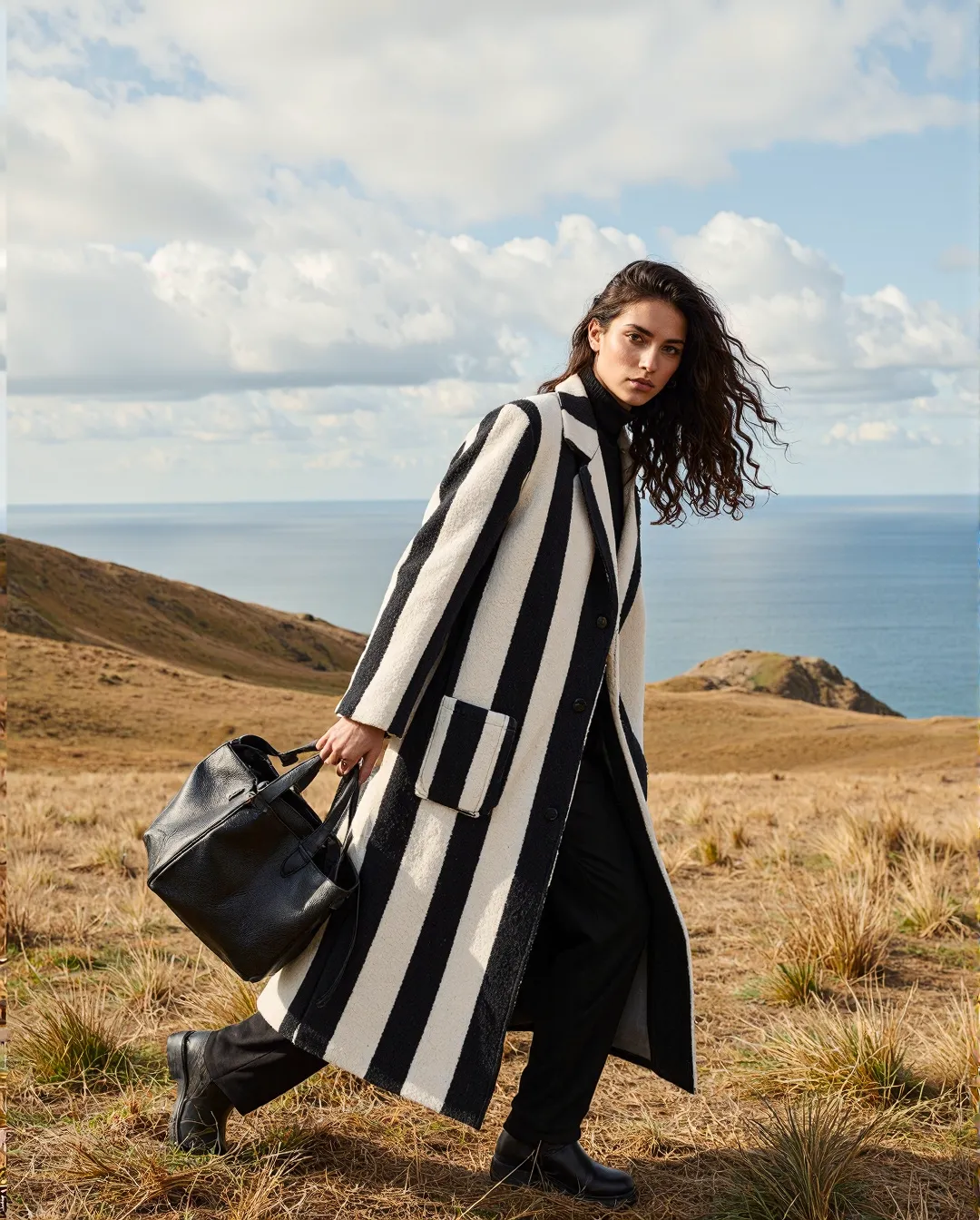 Striking black and white striped coat ensemble photographed against coastal hillside with ocean backdrop and dramatic cloudy sky