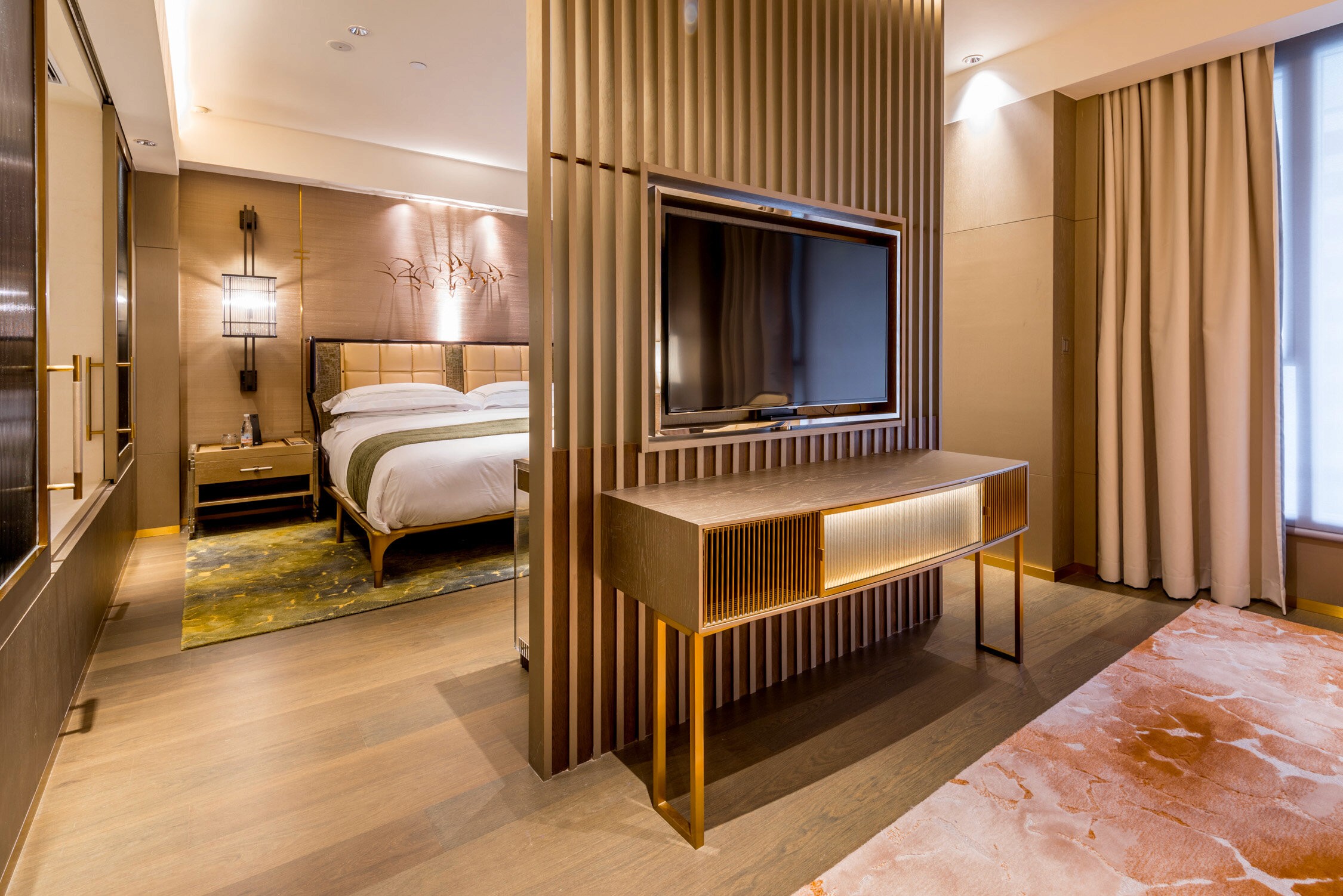 A modern hotel room features a large bed with white linens and a flat-screen TV mounted on a wood-paneled wall.