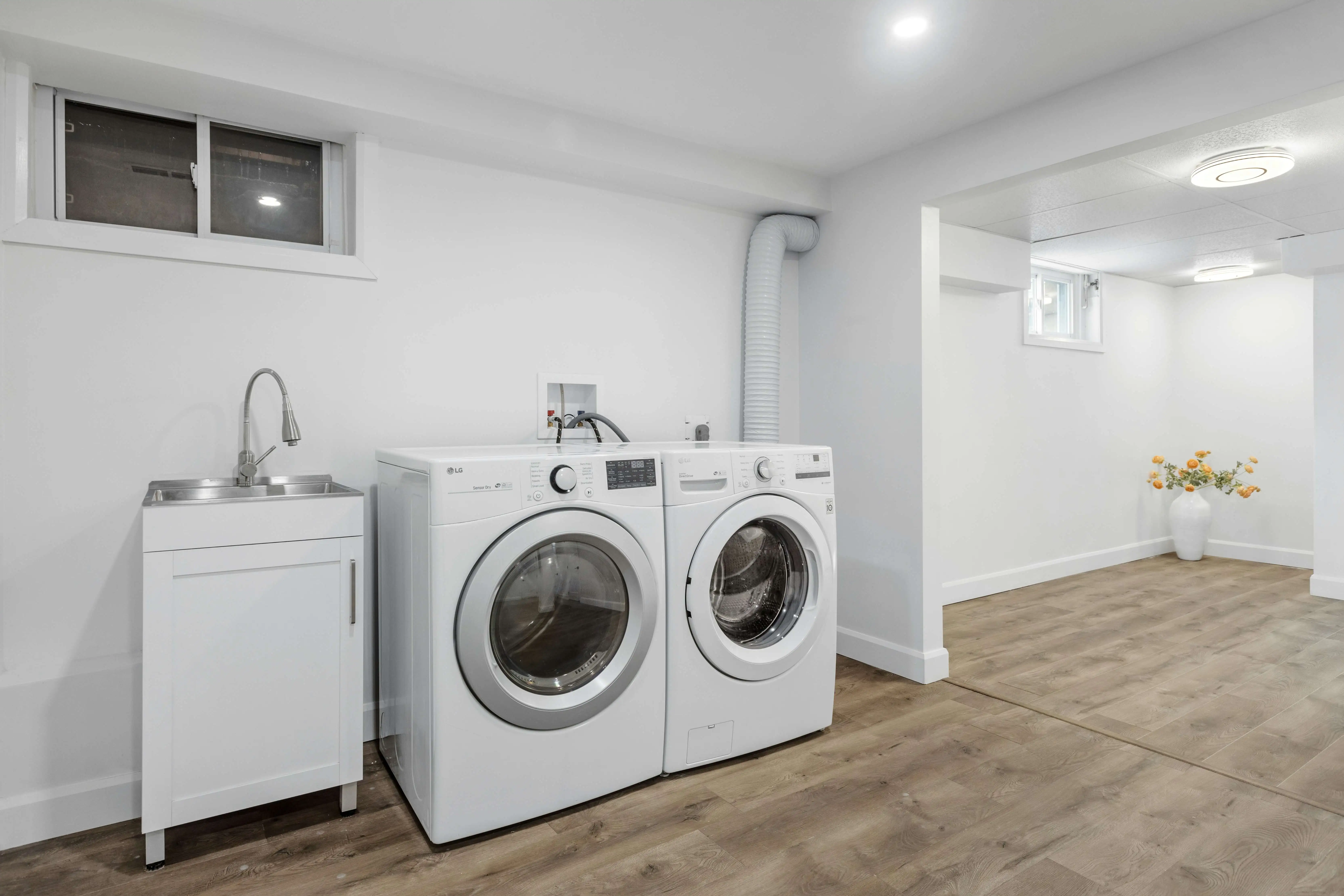 Modern laundry room with white washer and dryer next to a sink on wooden floor. Minimalist decor with a vase of flowers in the corner.