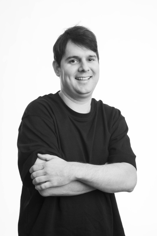 Black-and-white studio portrait of a smiling man with short dark hair, wearing a black t-shirt, arms crossed, against a plain light background.
